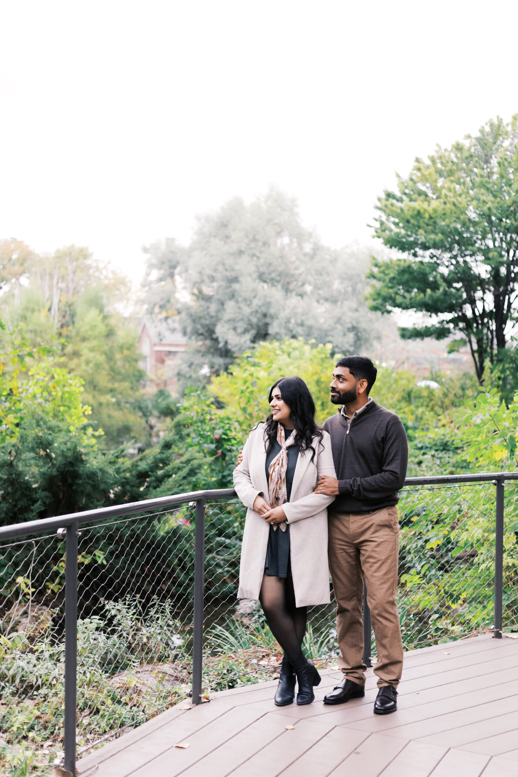 A couple stands on a wooden platform overlooking a garden, with trees and distant rooftops in the background.