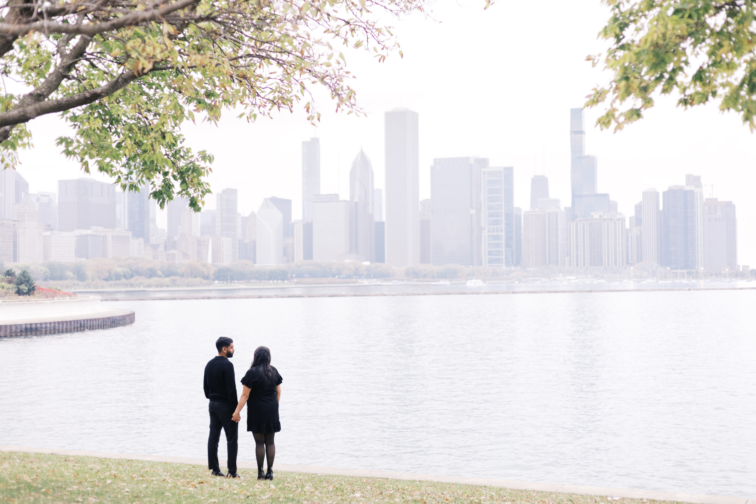 A couple holds hands while standing by the lakefront, looking out at the Chicago skyline under a cloudy sky.