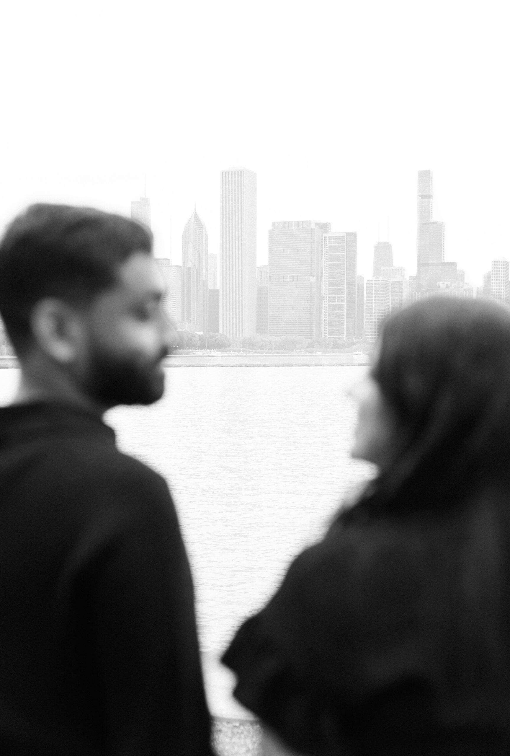 Black and white photo of a couple out of focus in the foreground, with the Chicago skyline clearly visible in the distance.