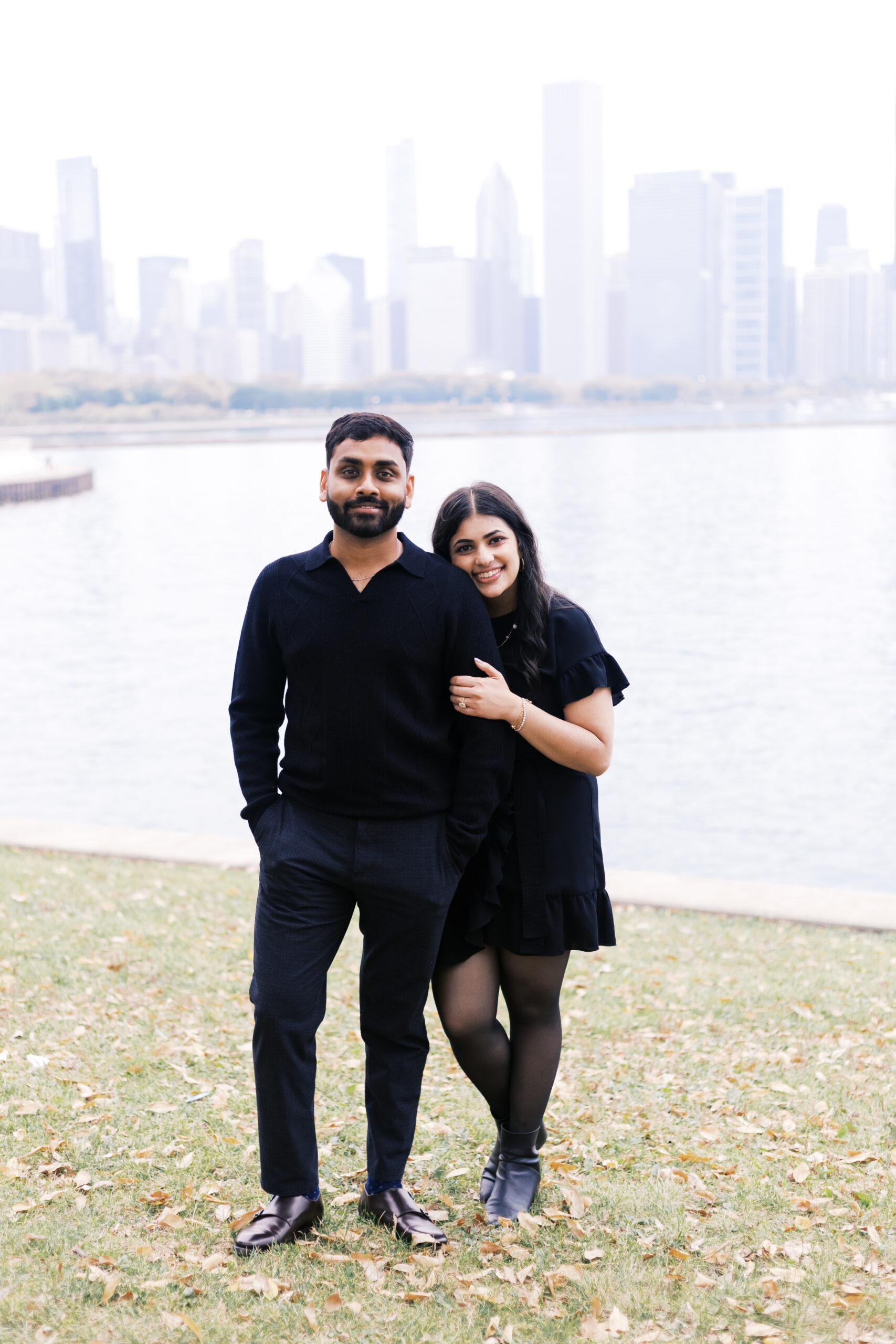 A woman leans against a man’s arm, both smiling, standing on grass by Lake Michigan with the city skyline behind them.