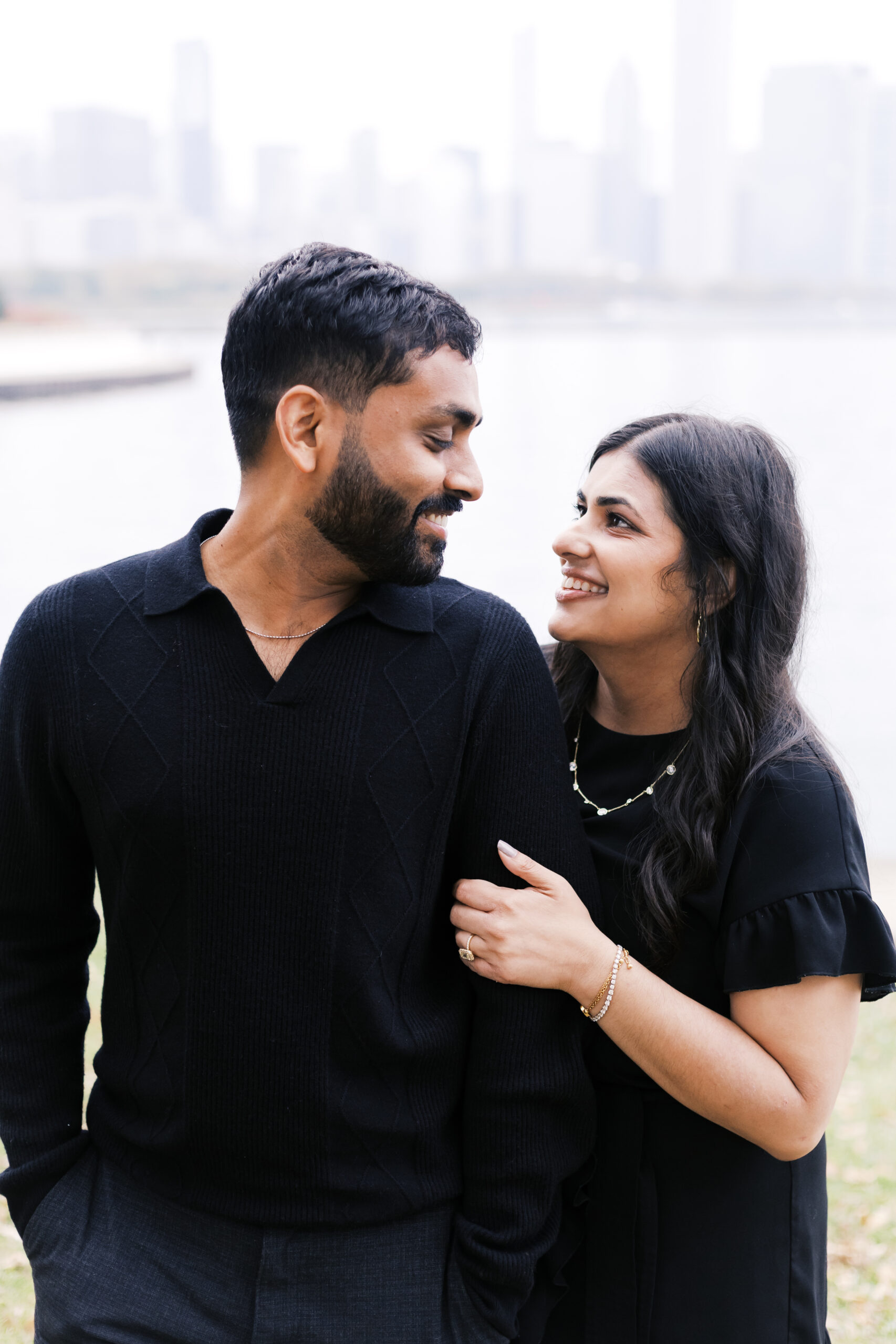 A couple smiles at each other near the lakefront, the woman holding the man's arm, with the city skyline in the background.