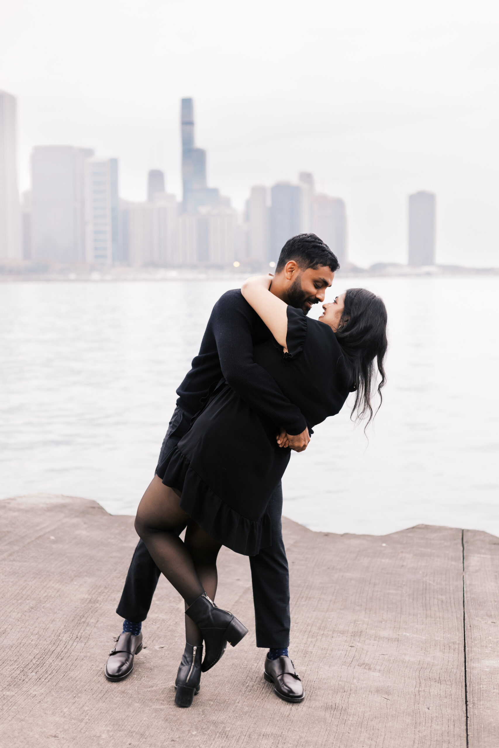 A man dips a woman in a playful pose on a concrete pier, both wearing black, with the Chicago skyline in soft focus behind them.
