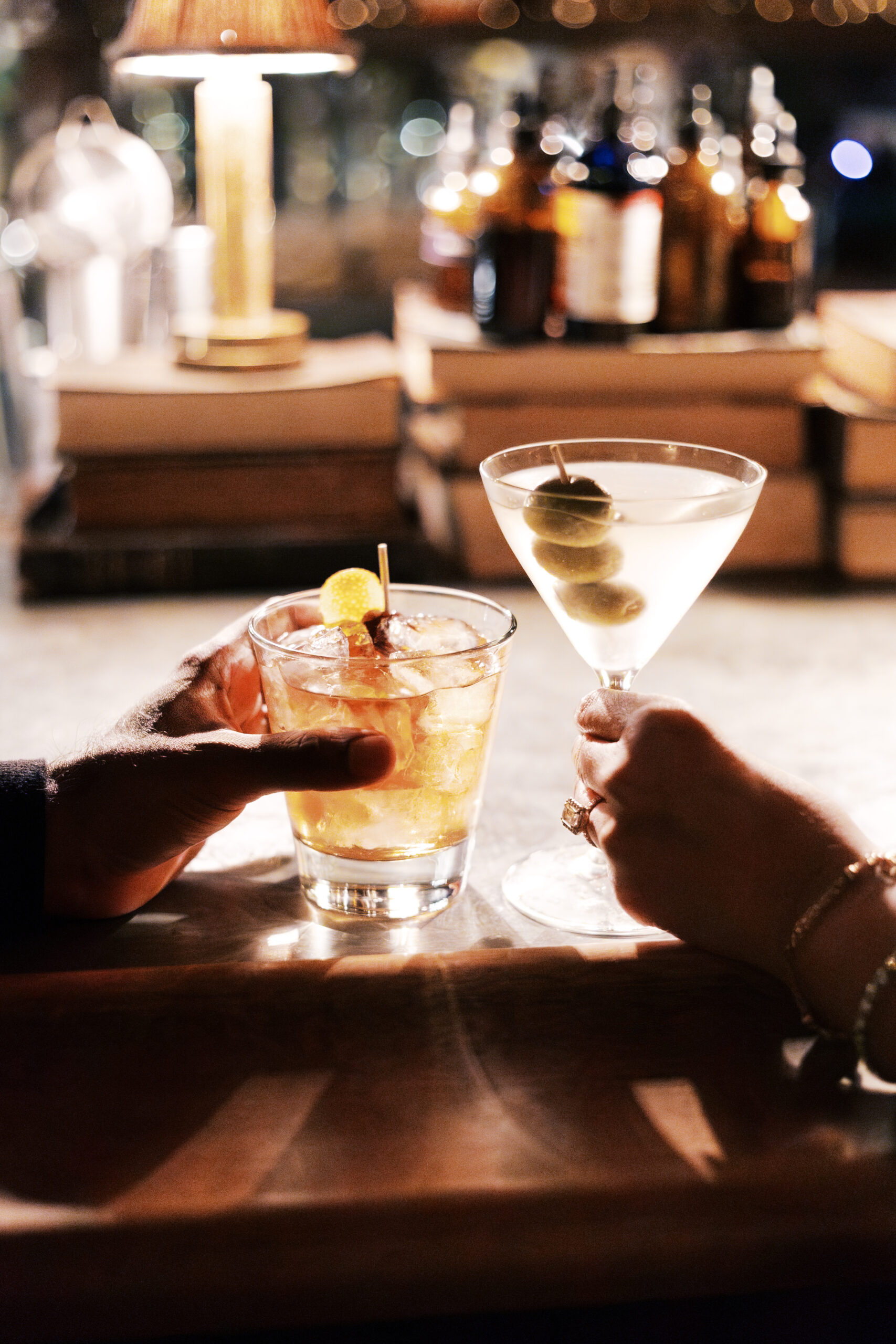 A close-up of two hands holding cocktails at a dimly lit bar—an Old Fashioned and a martini with olives.