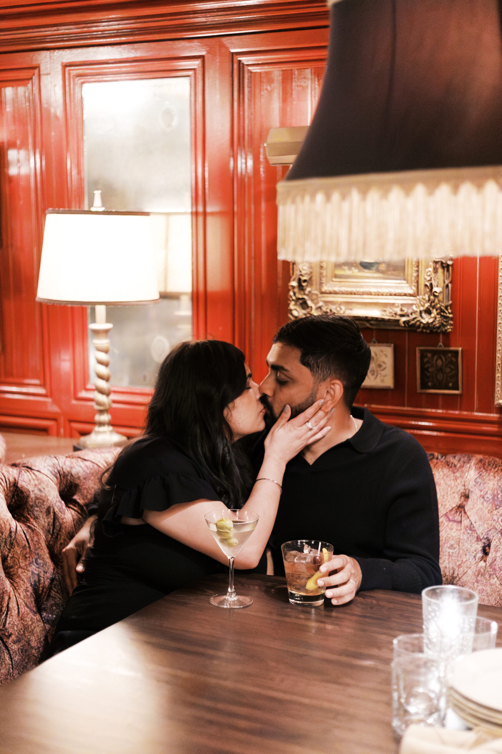 A woman gently kisses a man in a red-walled lounge as they sit close together with drinks on the table.