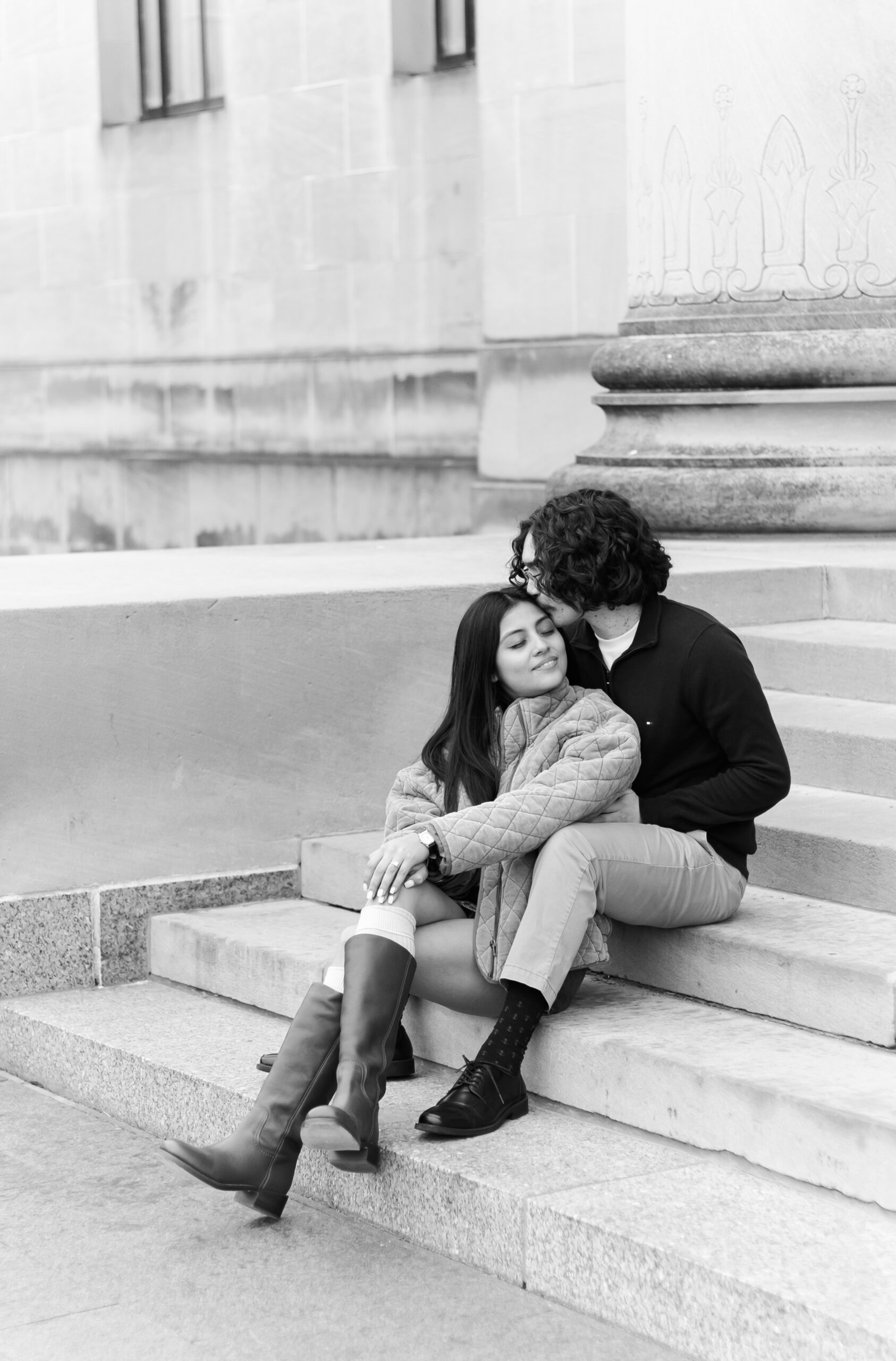 Black and white photo of a couple sitting on stone steps, one person kissing the other's temple while they lean in close.