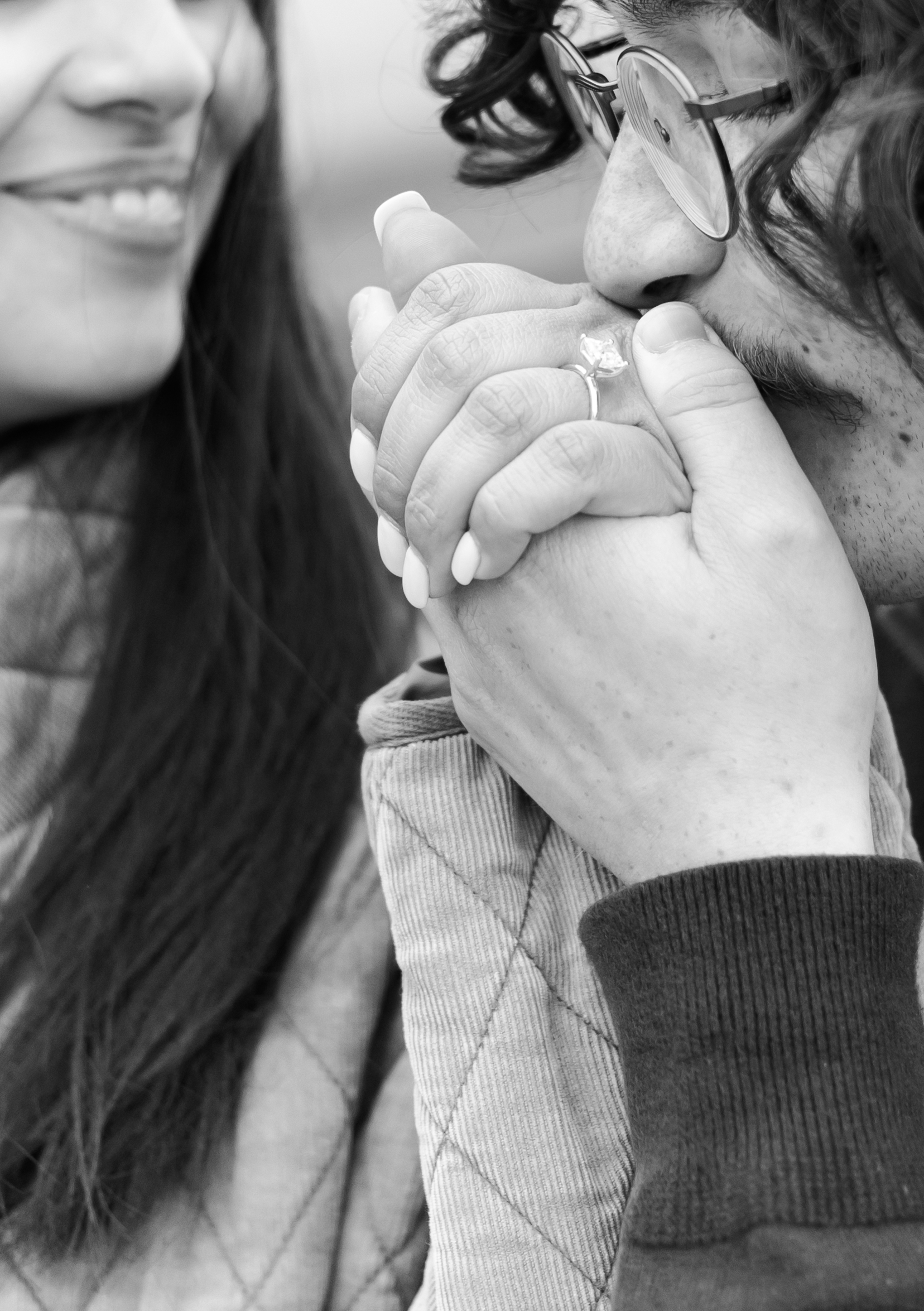 Black and white close-up of a person kissing their partner’s hand, highlighting the pear-shaped engagement ring.