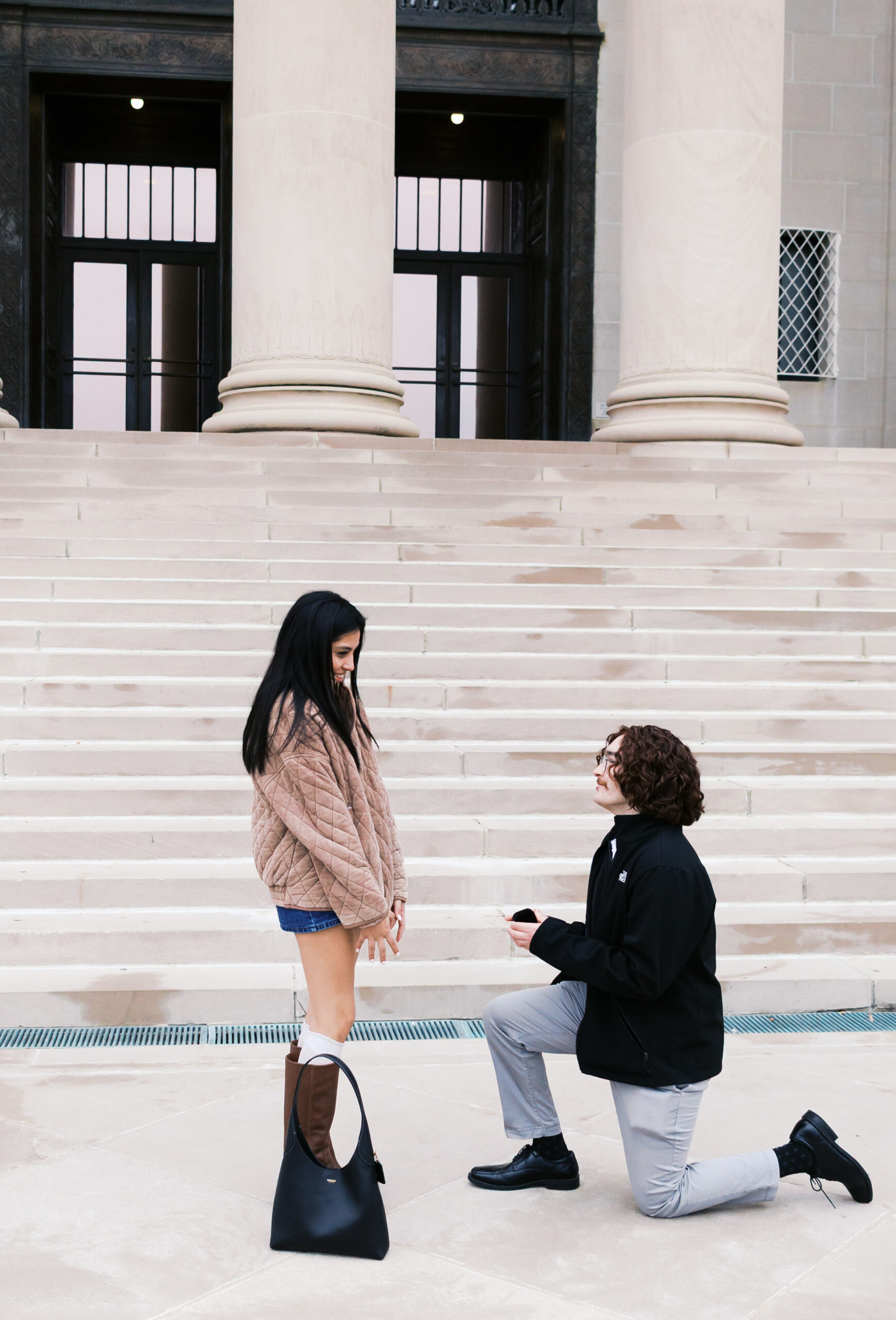 Person kneeling to propose to their partner on stone steps in front of a grand building with tall columns.