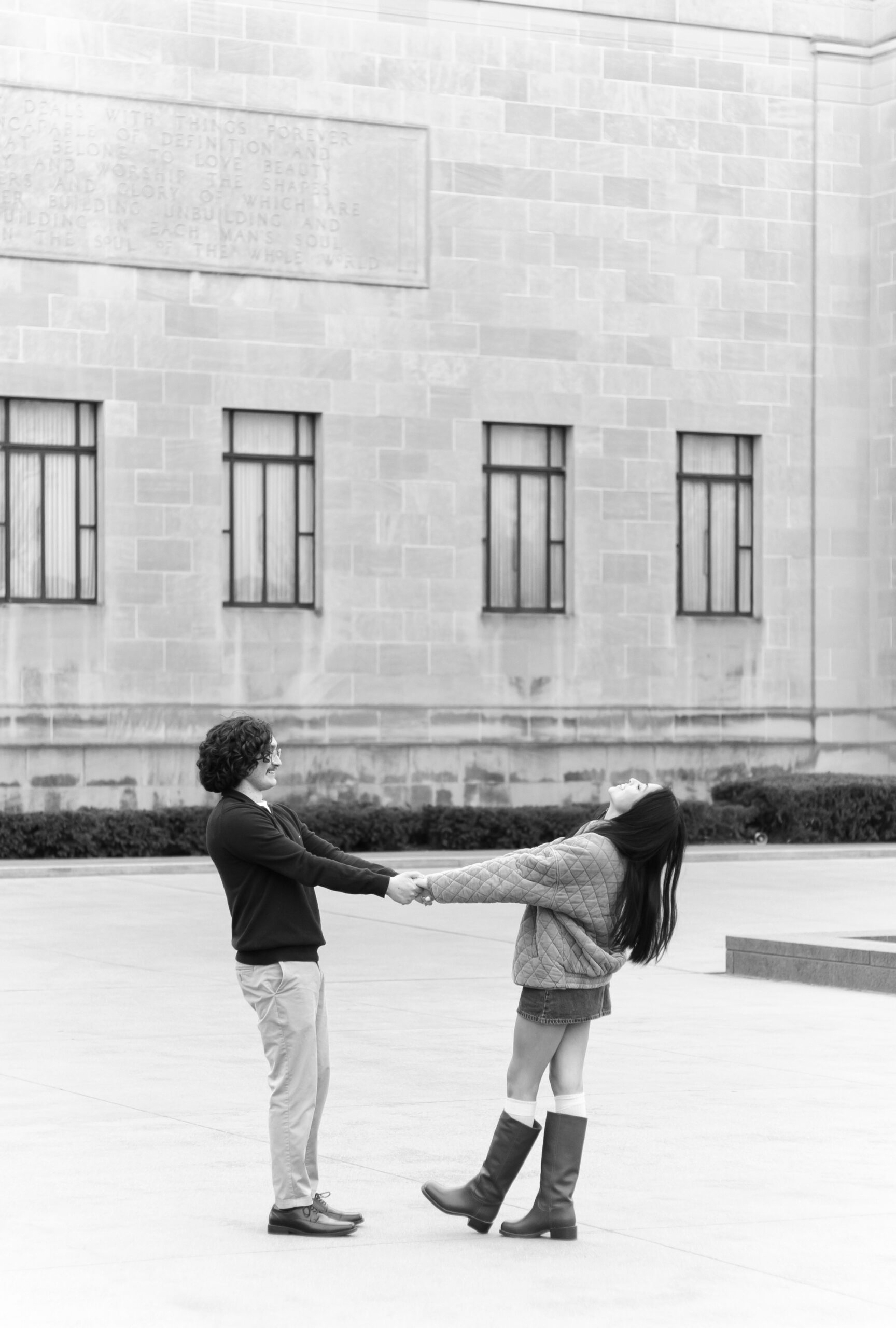 Black and white photo of a couple holding hands and playfully leaning back in an open courtyard.