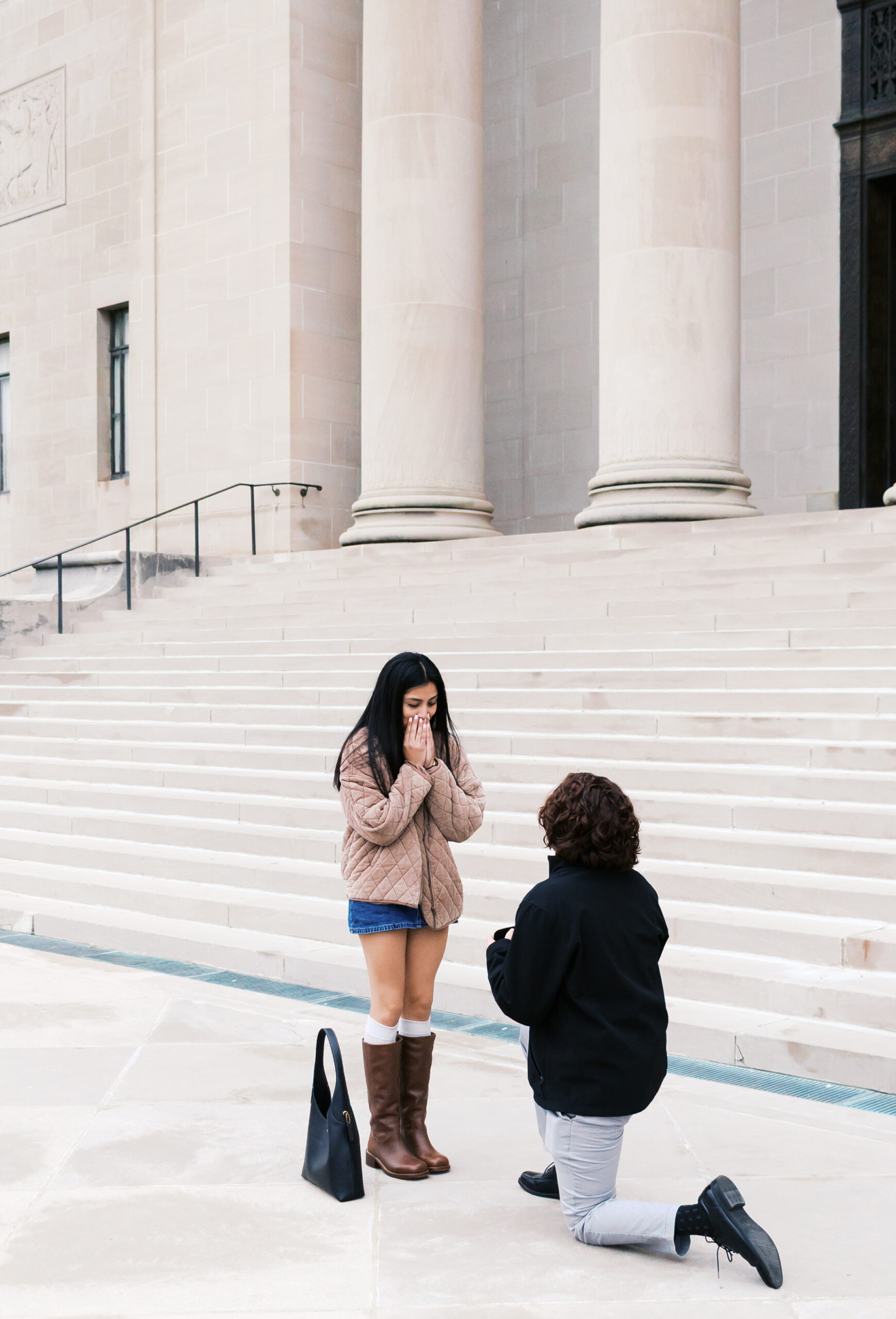 Person covering their mouth in surprise as their partner proposes on one knee in front of a historic building.