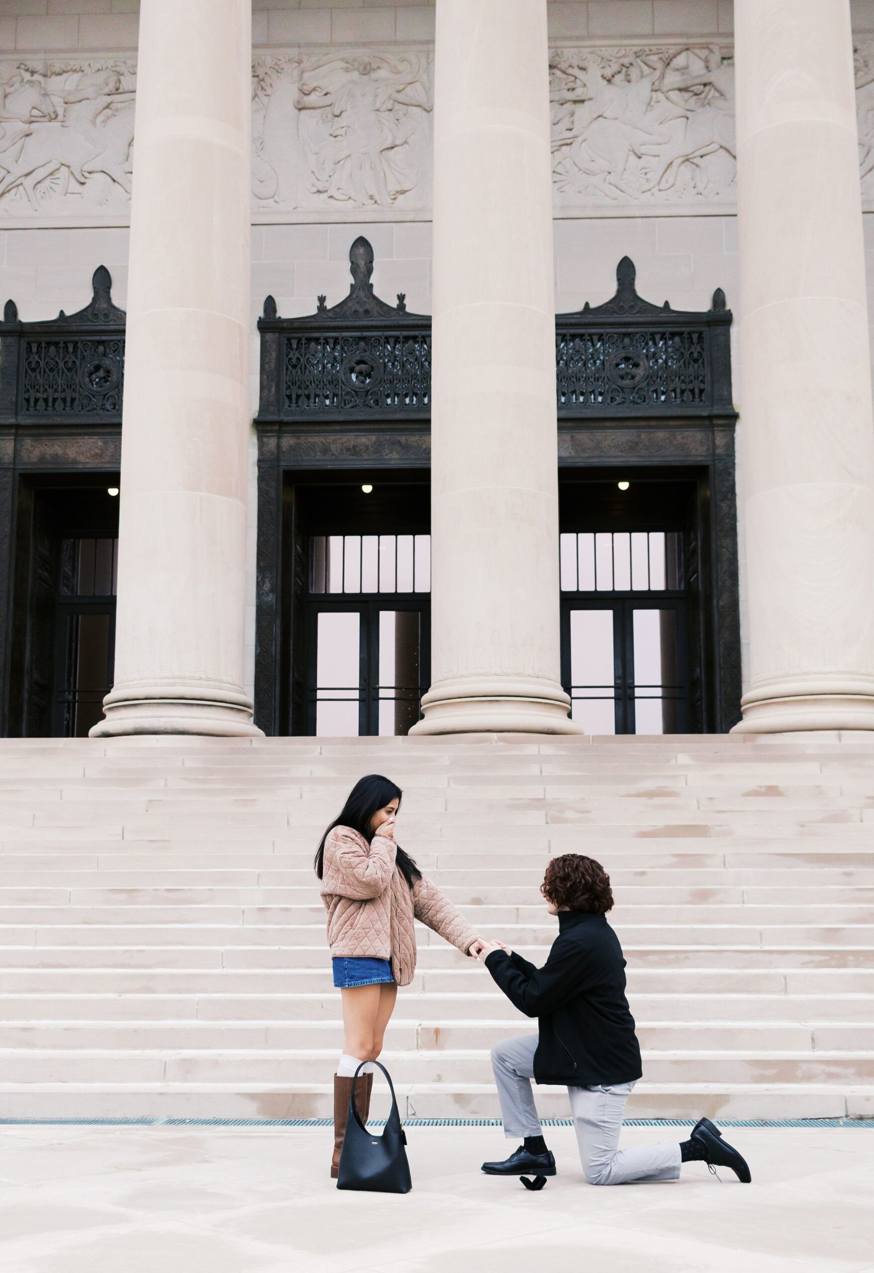 Person on one knee placing a ring on their partner’s hand during a proposal in front of tall columns.