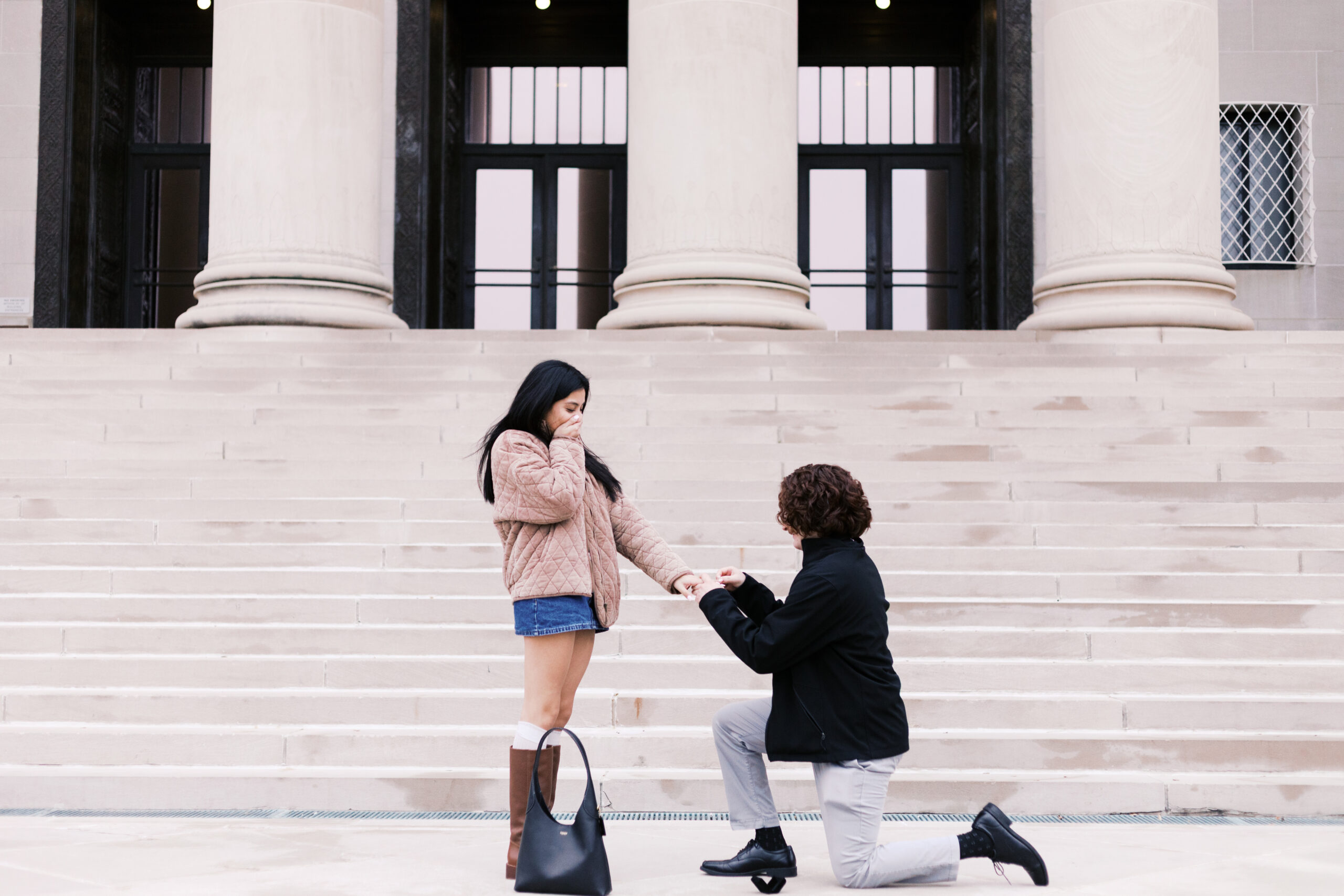 Couple during a proposal, one person kneeling and holding the other's hand while they react emotionally.
