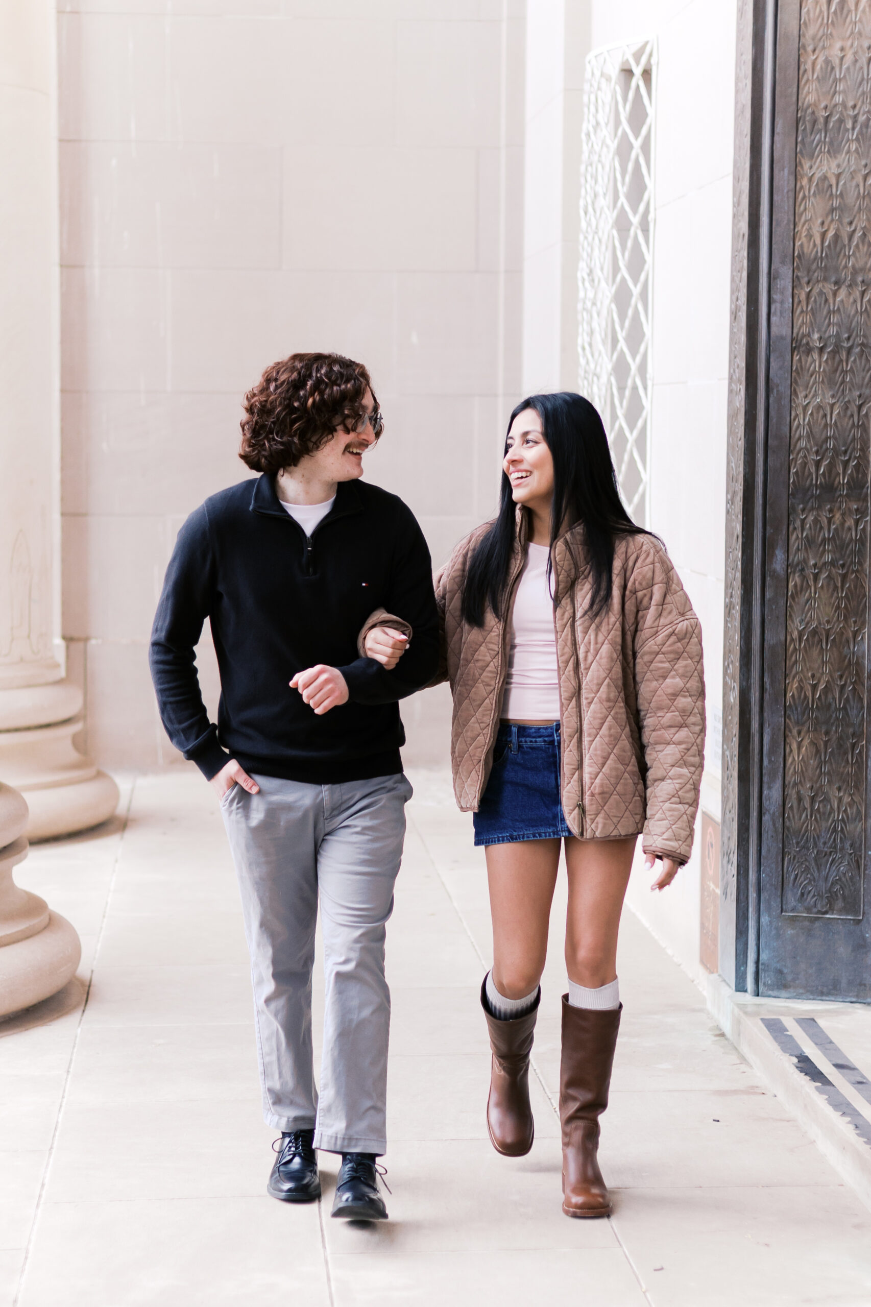 Couple walking arm in arm under a stone archway, smiling at each other after their engagement.