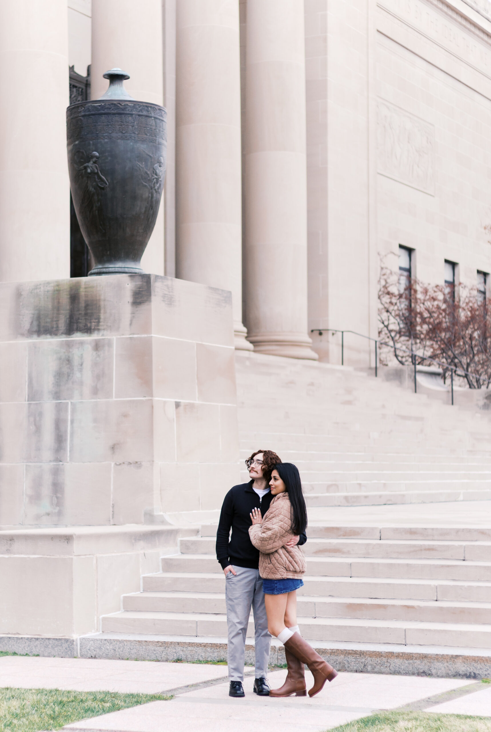 Couple standing together outside a neoclassical building, the partner with the ring resting a hand on the other's chest.