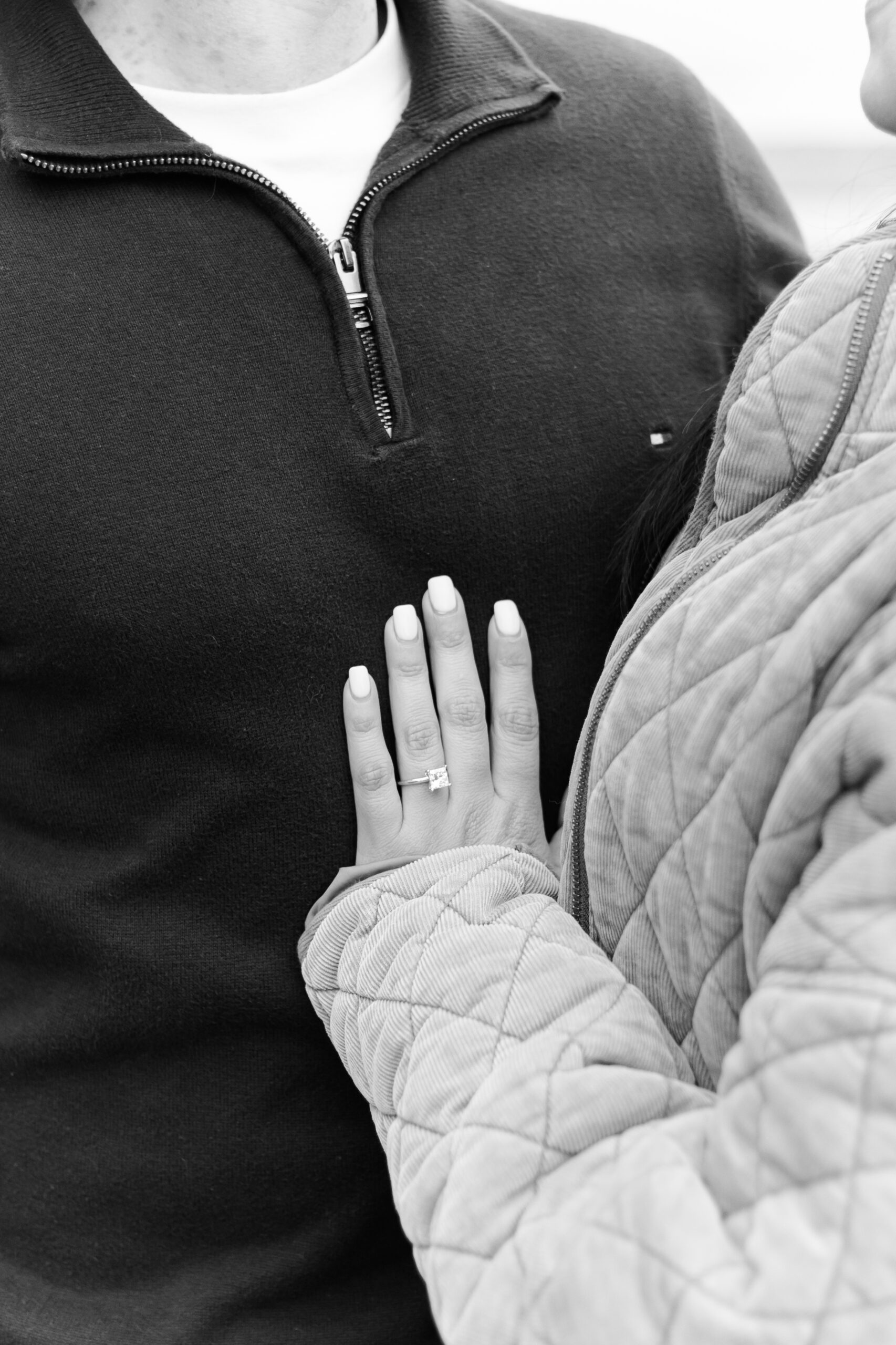 Black and white close-up of an engagement ring on a person’s hand resting against their partner’s chest.