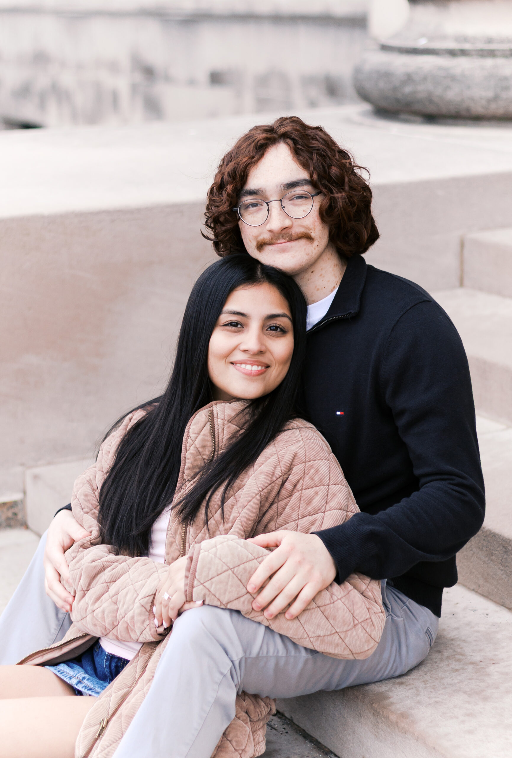Couple sitting on stone steps, embracing with smiles after their engagement, the ring visible on the person’s hand.