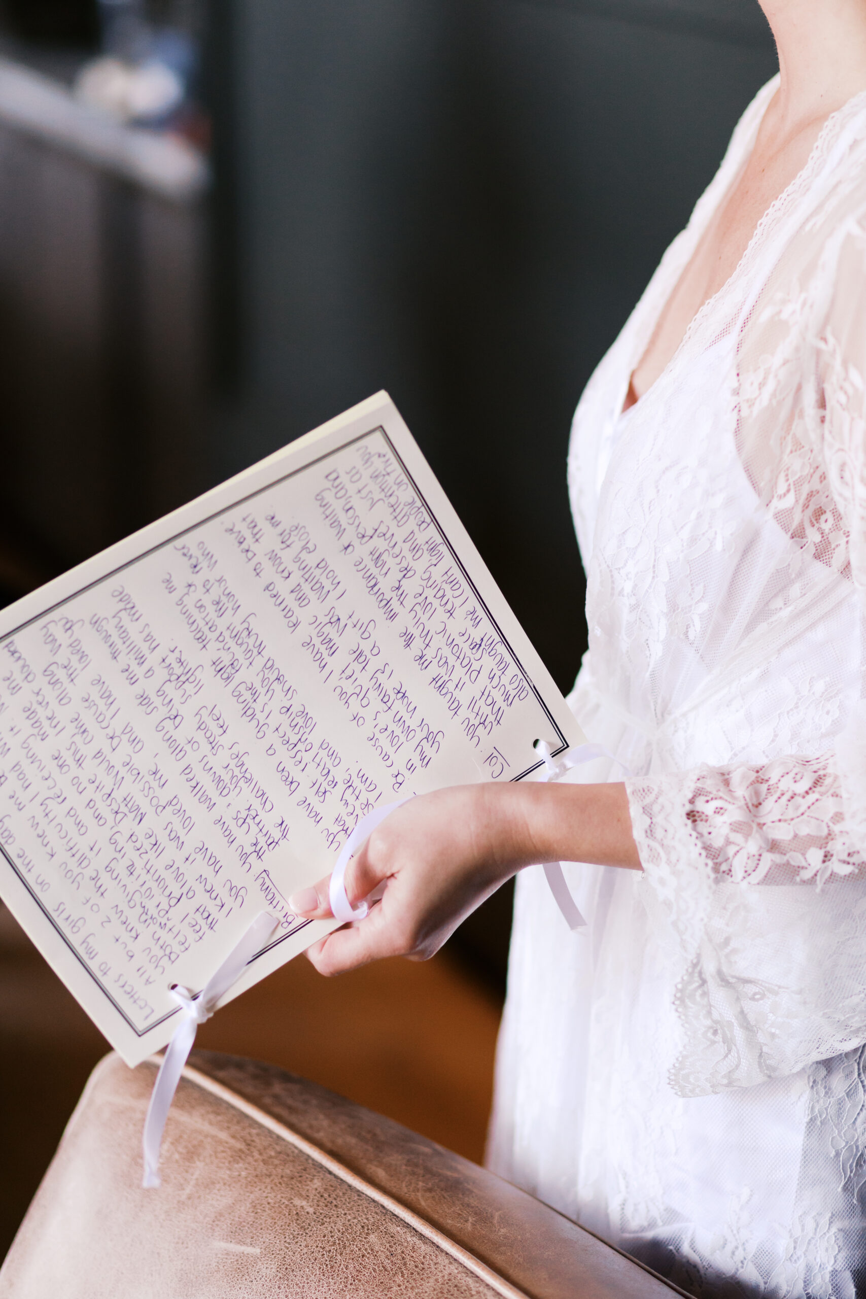 Bride in lace robe holding a handwritten wedding vow booklet tied with ribbon on wedding day morning.