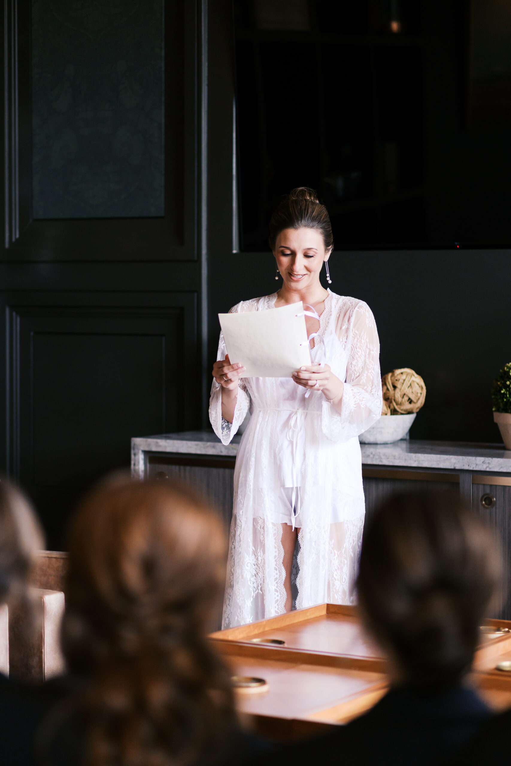 Bride in a white lace robe reading her vows aloud to her bridal party.