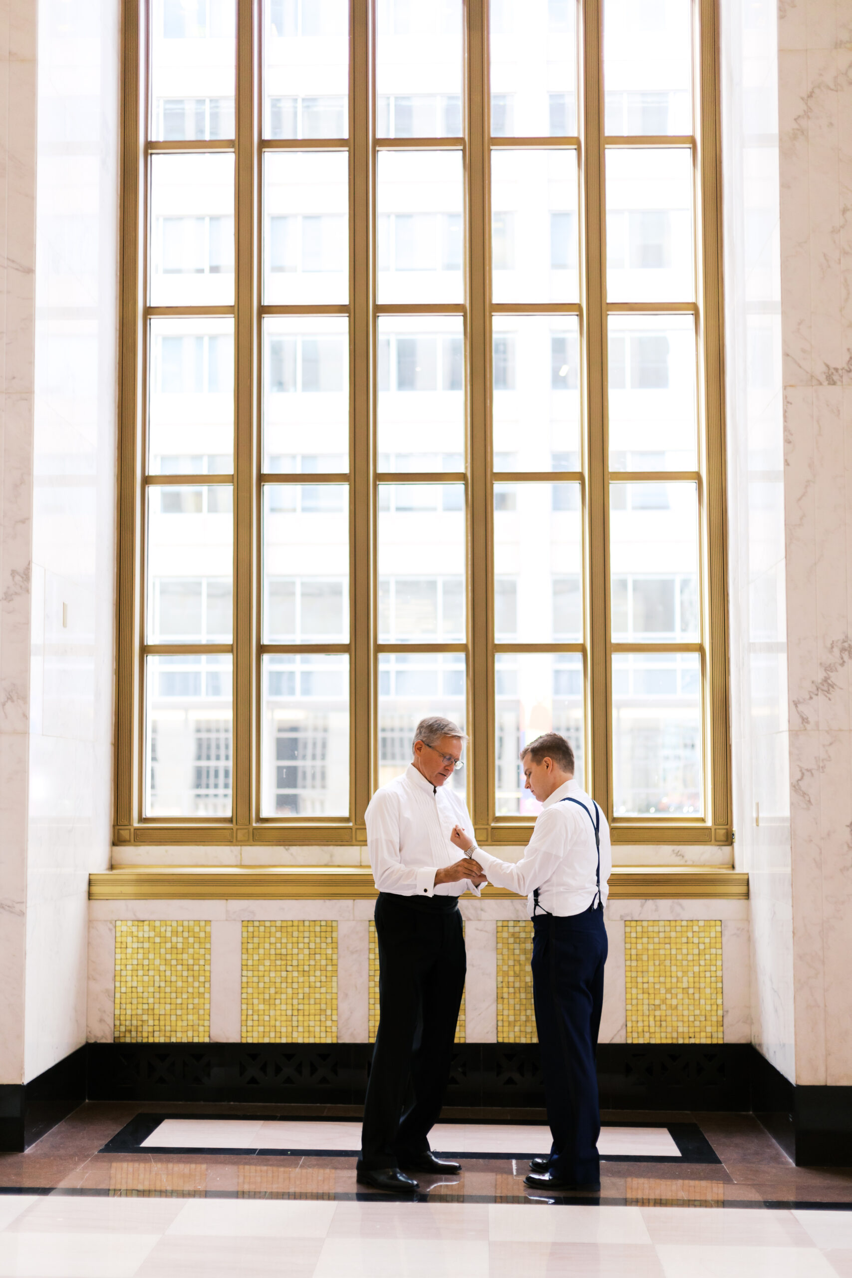 Groom and father in formalwear adjusting cufflinks by tall windows inside The Old Post Office in Chicago.