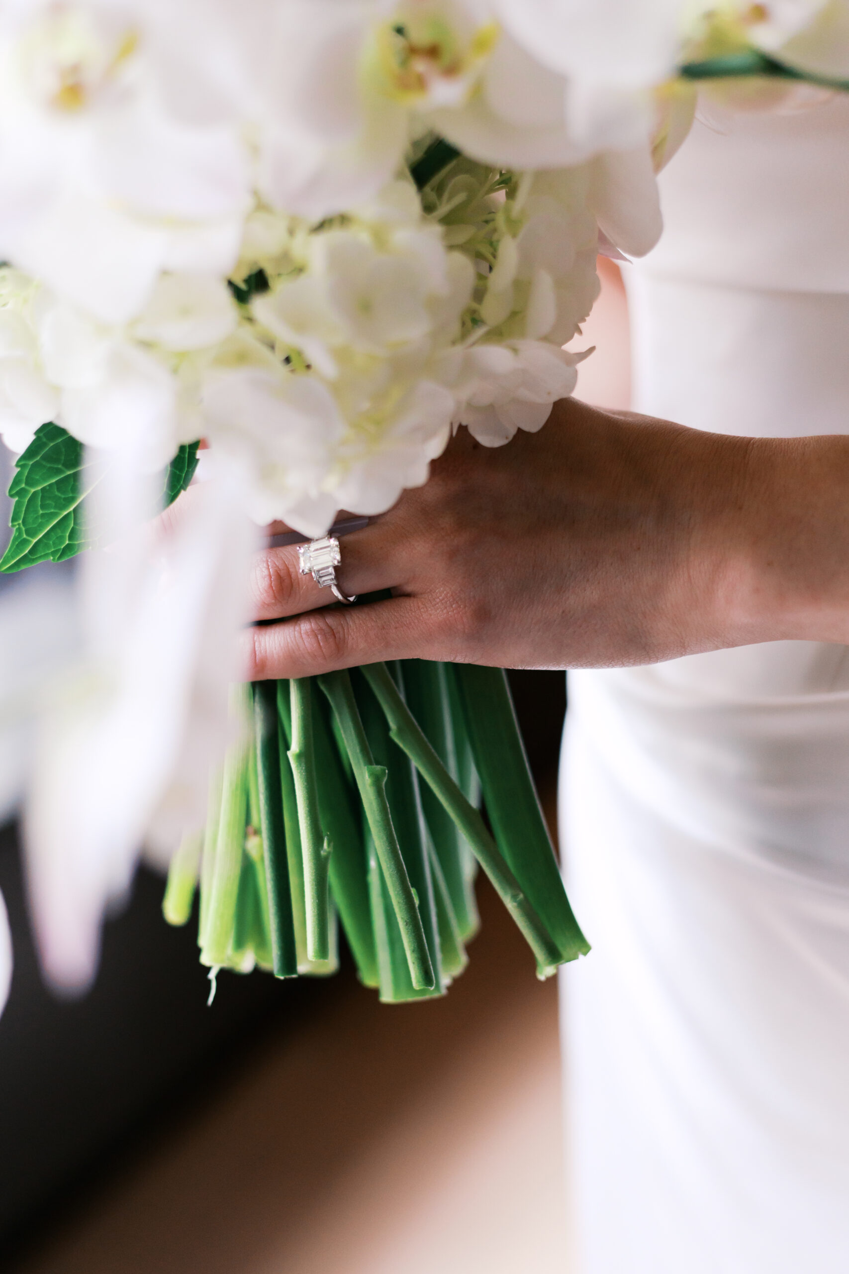Close-up of bride’s hand holding a white orchid and hydrangea bouquet, showing her emerald-cut engagement ring.