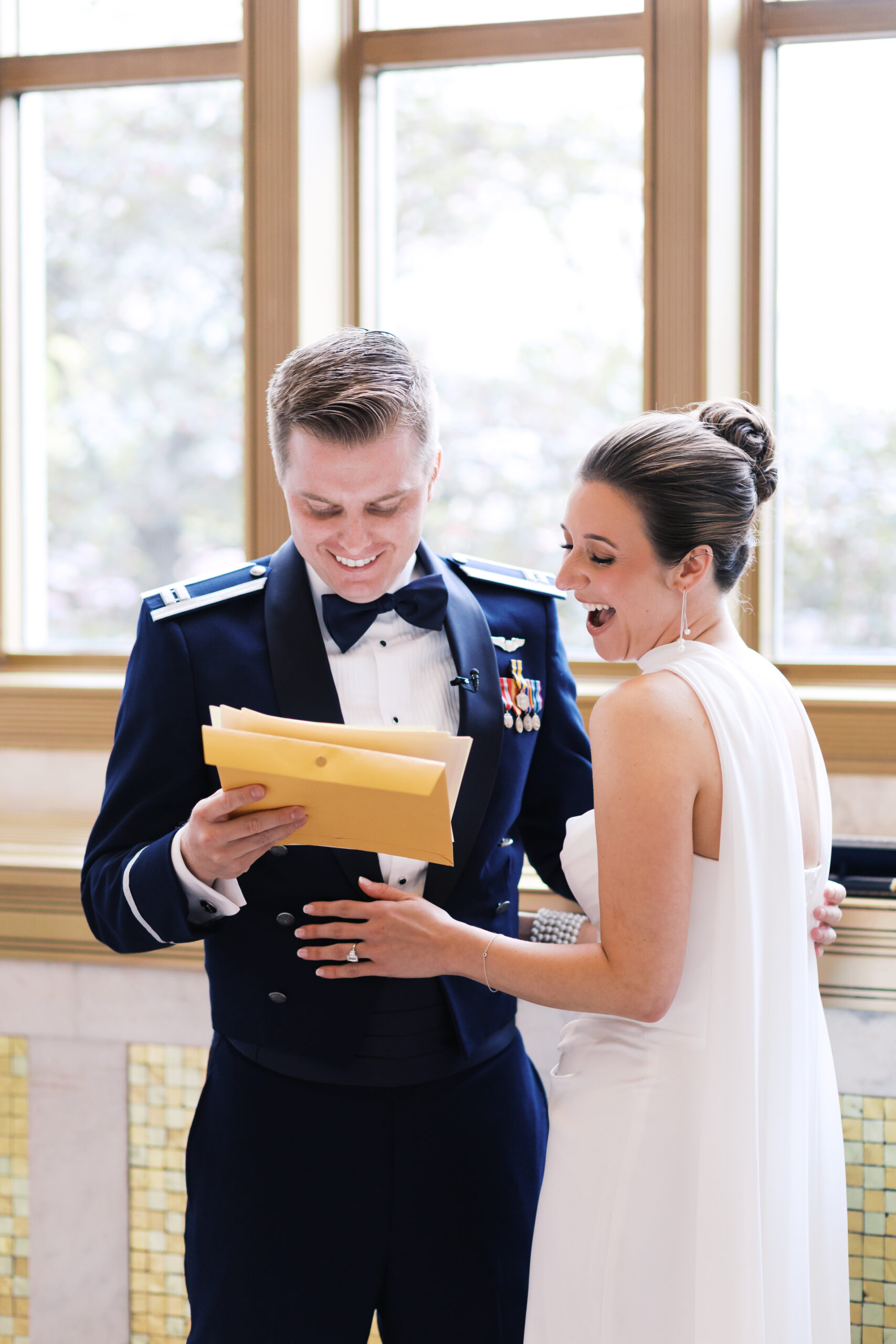 Bride and groom smiling as he reads a letter from her, standing by the windows.