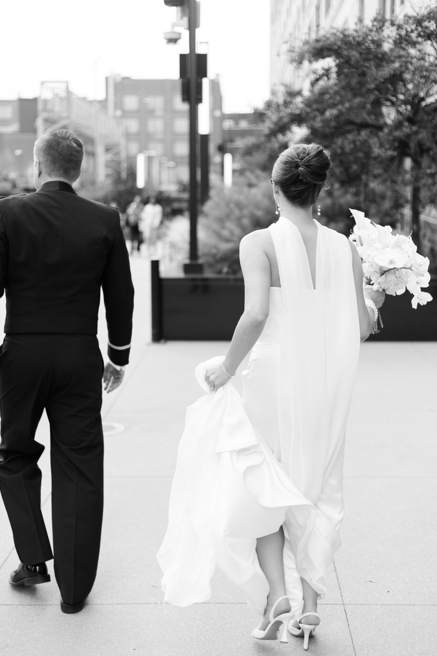 Black and white image of bride holding her gown and bouquet while walking beside the groom outdoors in downtown Chicago.
