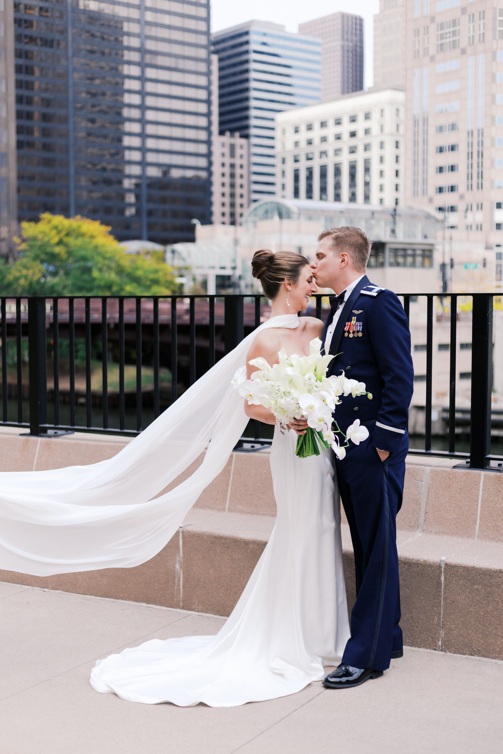 Groom in military dress uniform kisses bride on the forehead as she smiles, with the Chicago skyline in the background.