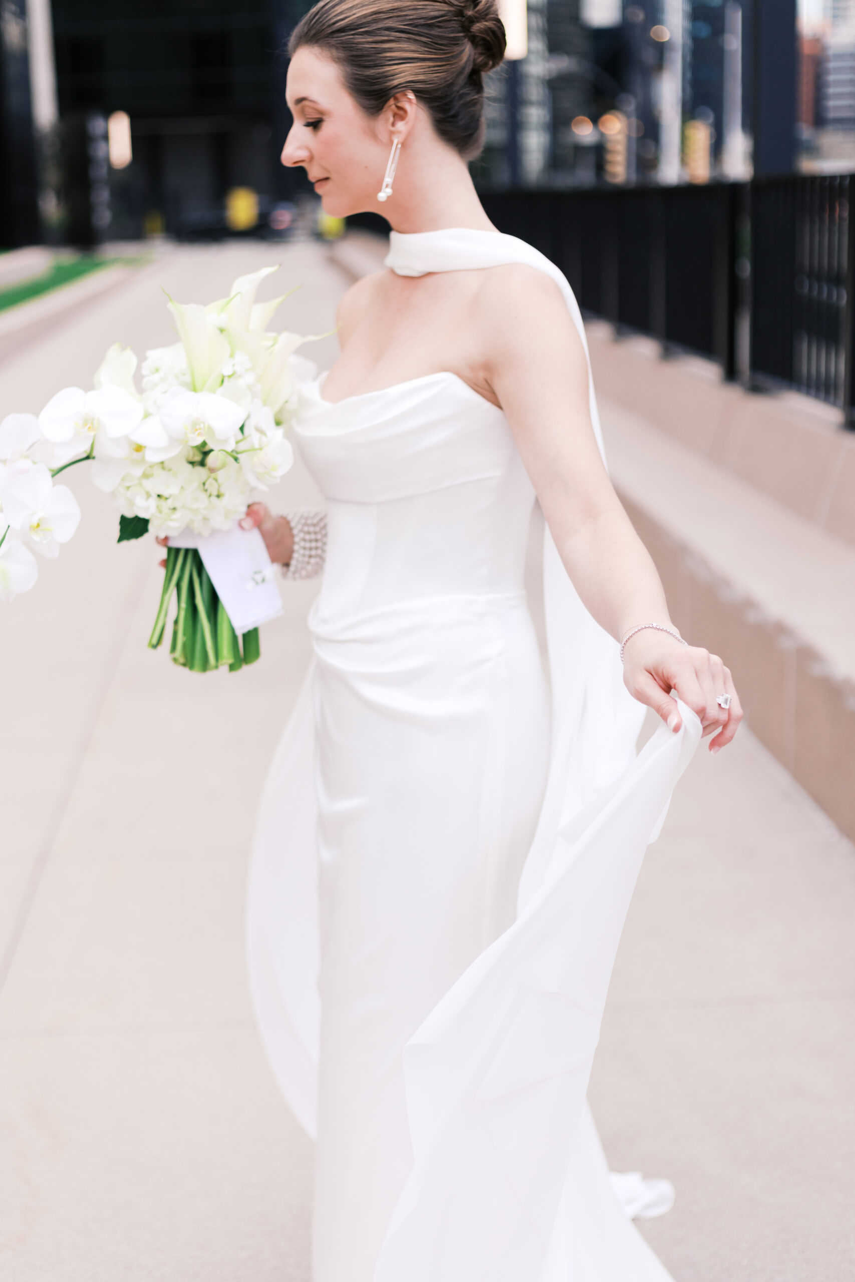 Bride in a satin gown holds her bouquet and gently lifts the hem of her dress while walking along a city sidewalk.