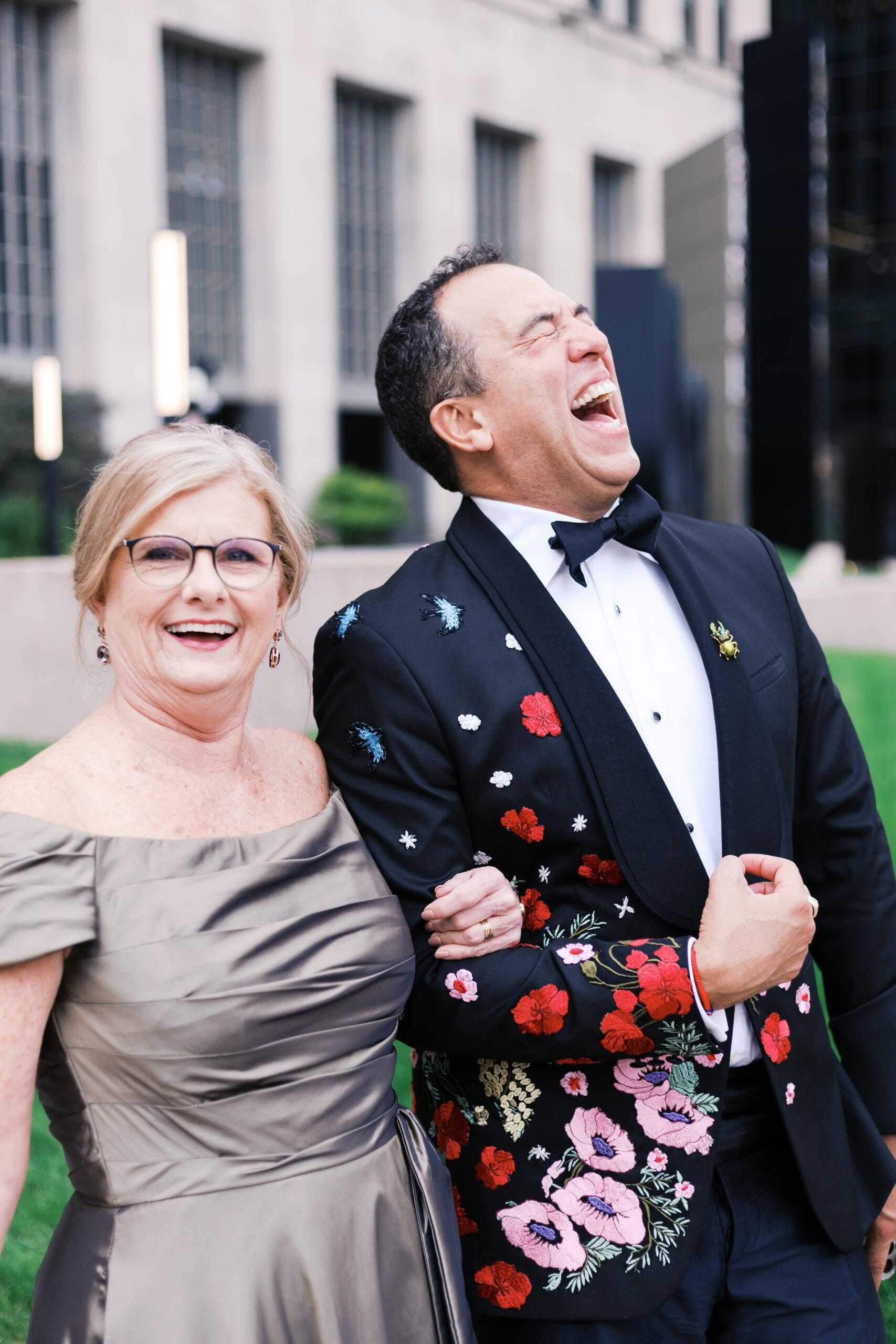 A man in a floral embroidered tuxedo laughs with a woman in an off-the-shoulder gown, walking arm in arm at the wedding.