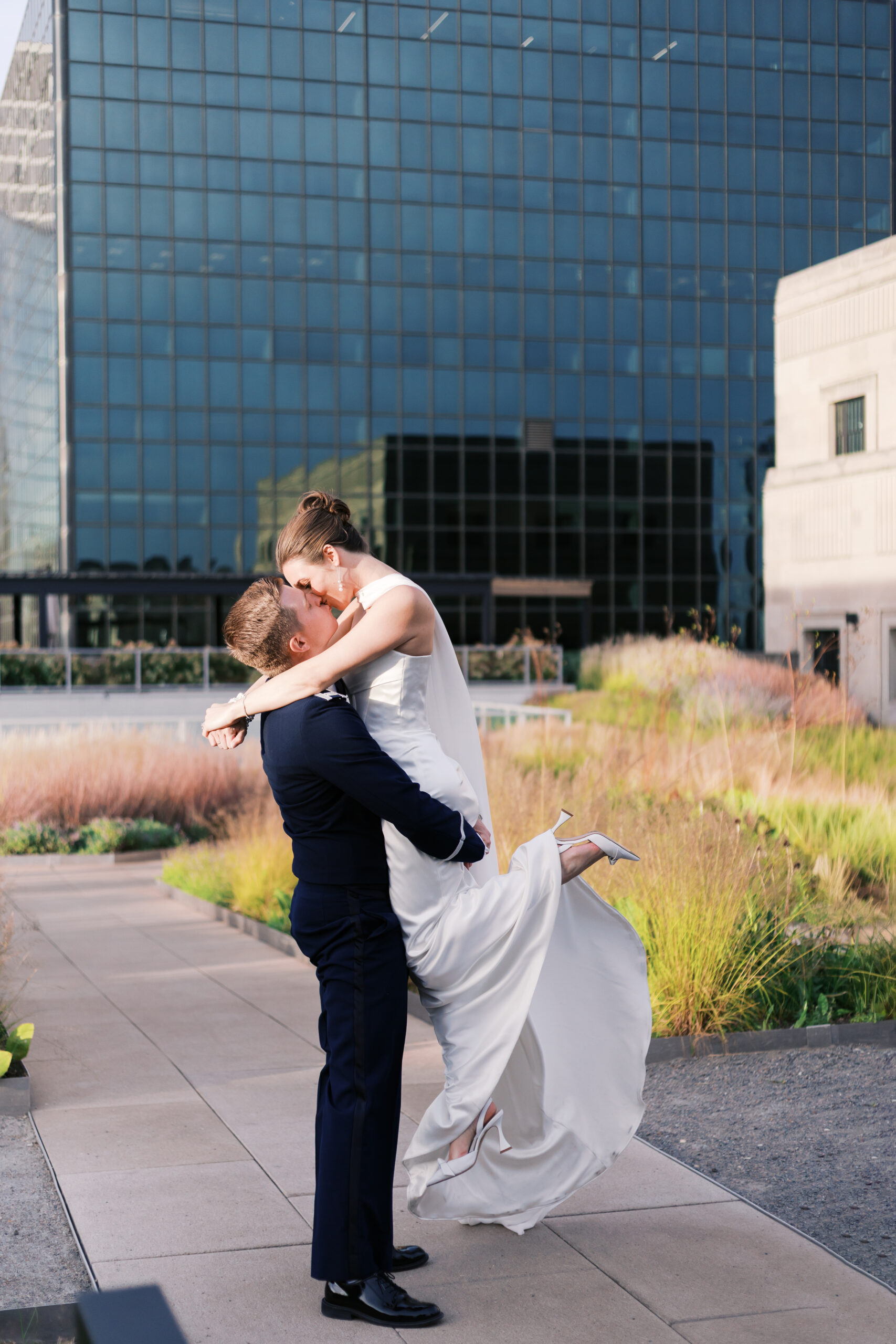 Groom lifts bride in an embrace as they kiss, her heels off the ground, surrounded by tall grasses on a rooftop in downtown Chicago.