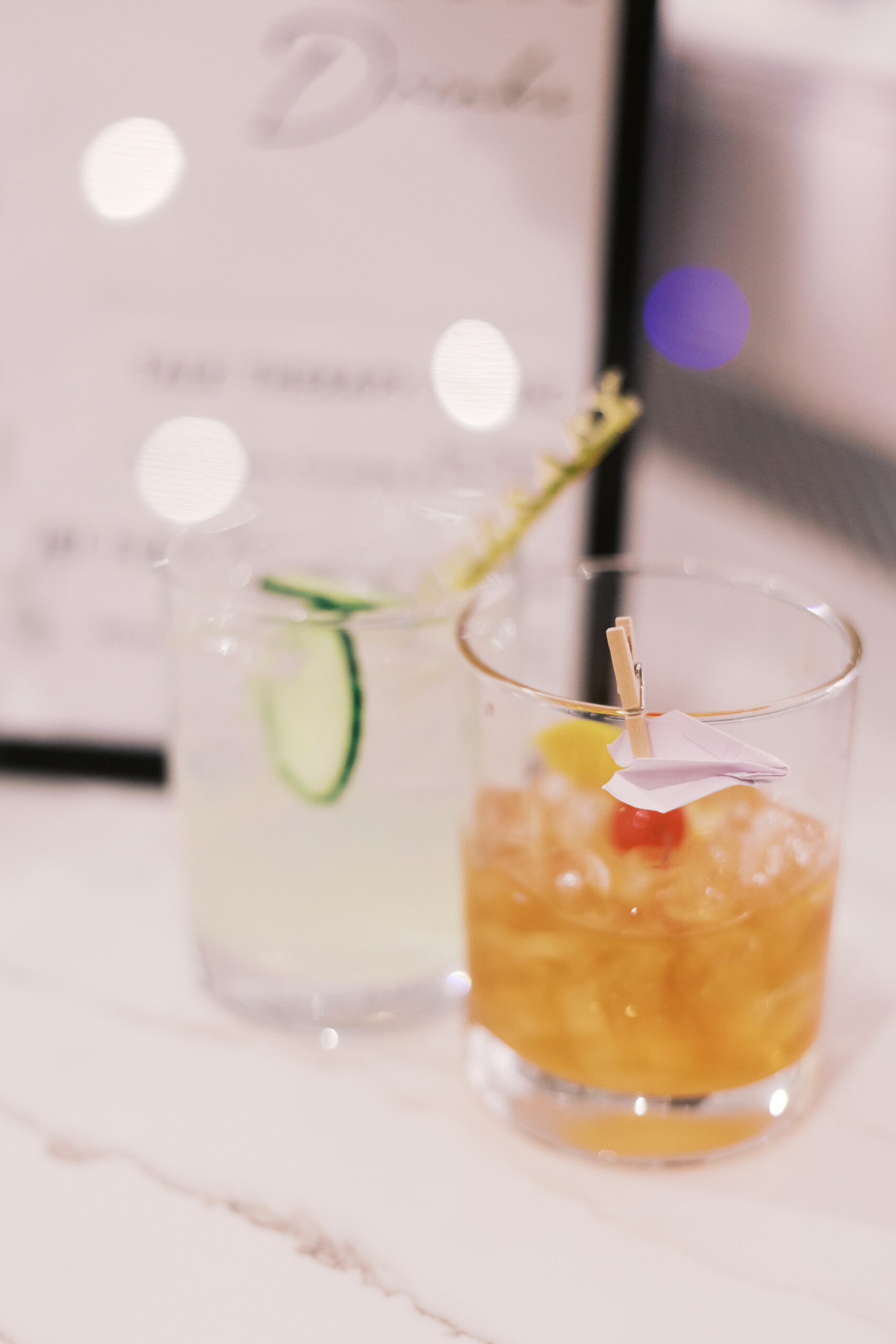 Two signature wedding cocktails on a marble bar, including a drink with a cucumber garnish and another with a paper airplane detail.