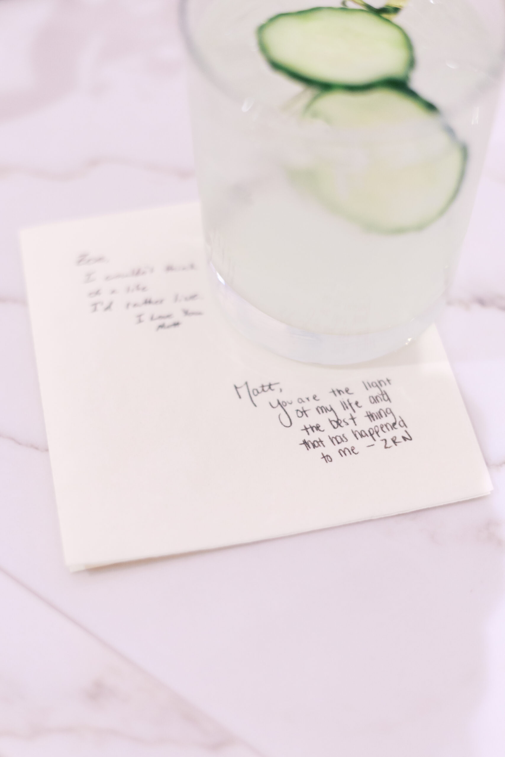Handwritten love notes resting under a cocktail glass with cucumber slices on a marble countertop.