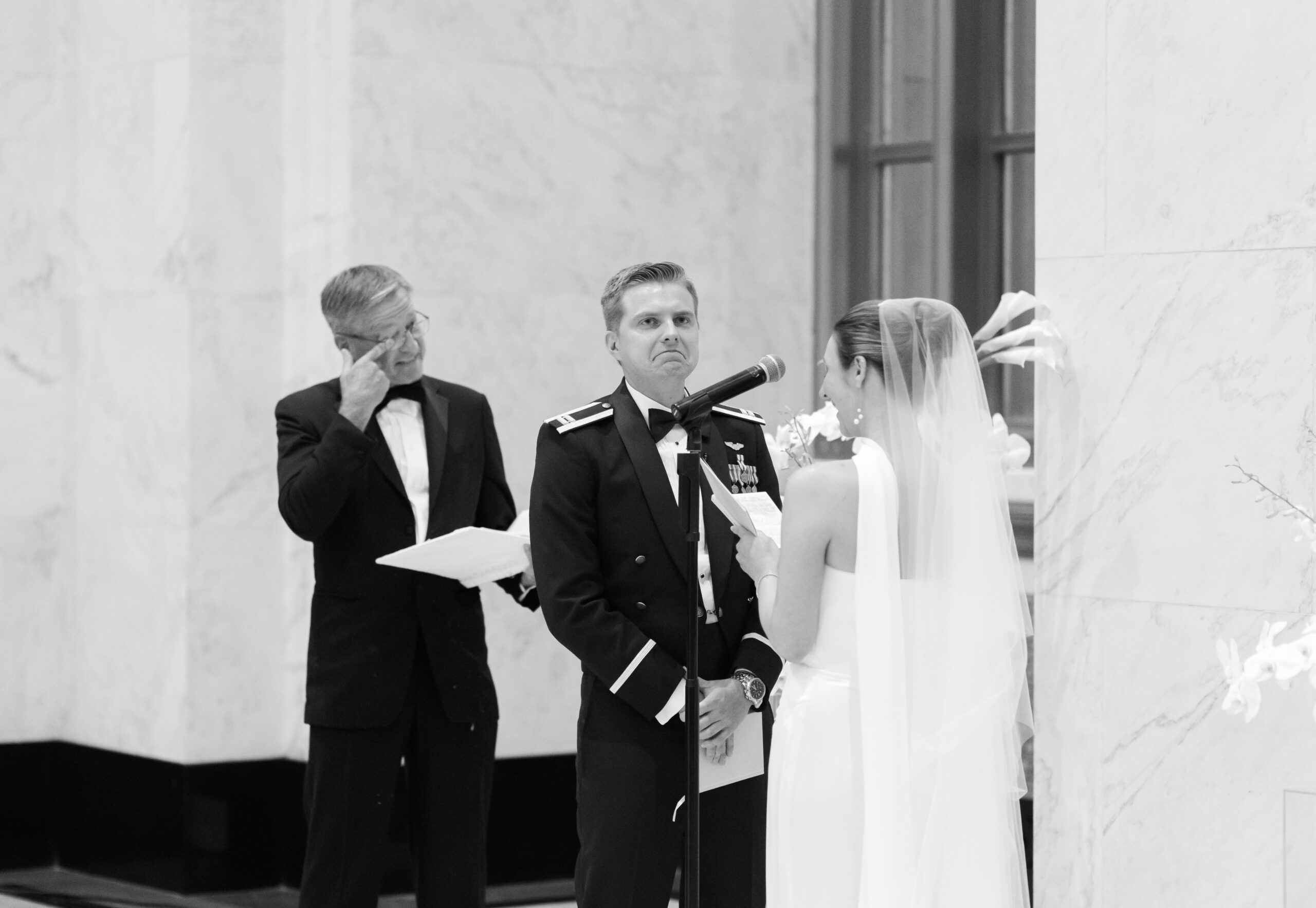 Father of the groom wipes away a tear as the bride reads her vows to the groom in a black and white image.