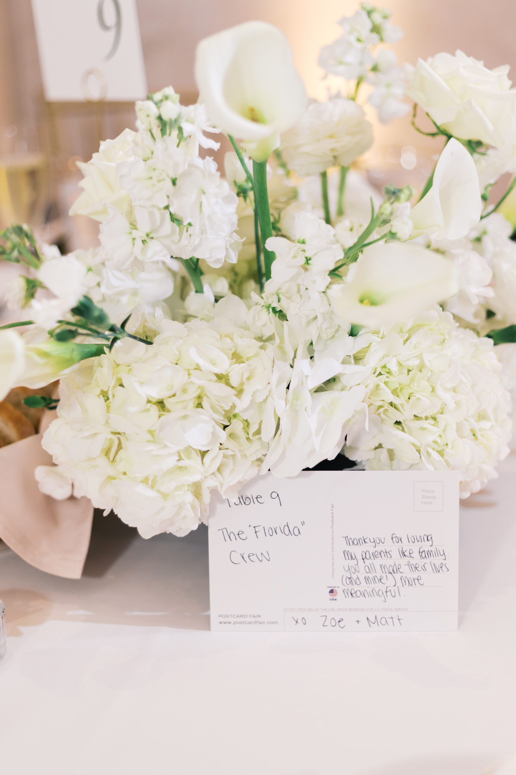 Close up of a floral arrangement with white calla lilies and white hydrangeas with a post card table number 9 titled the Florida Crew