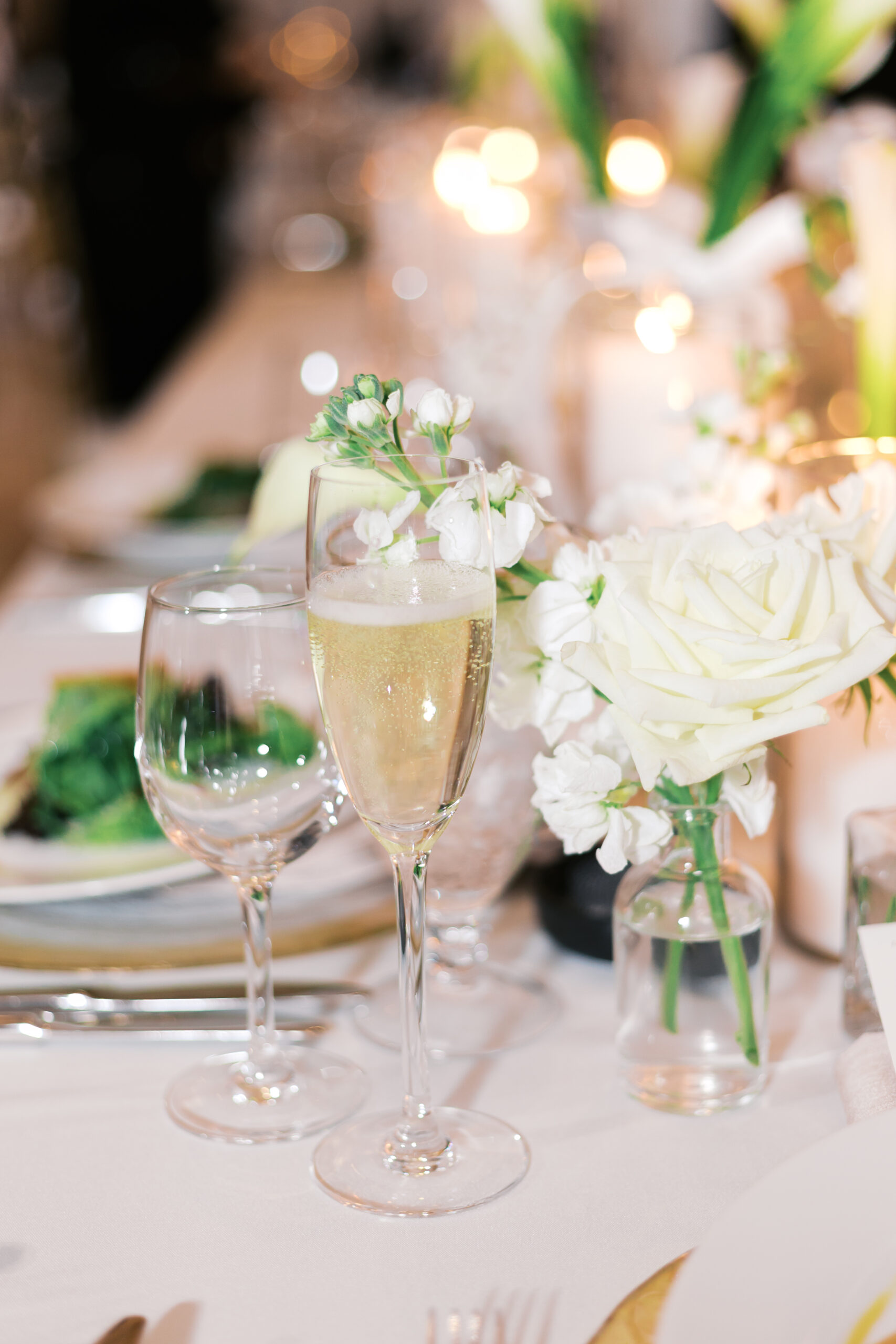 Glass of champagne on a reception table with a bud vase with a white rose nearby.