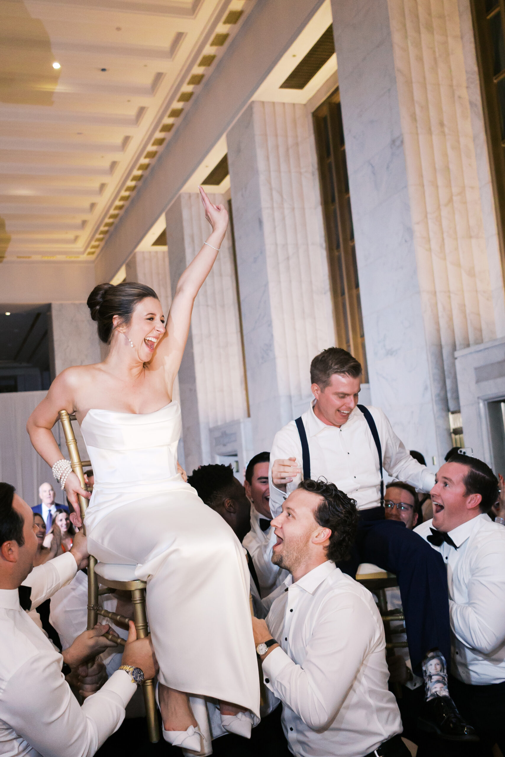 Bride raising her arm in excitement as she's raised in a chair on the dance floor with the groom in the background