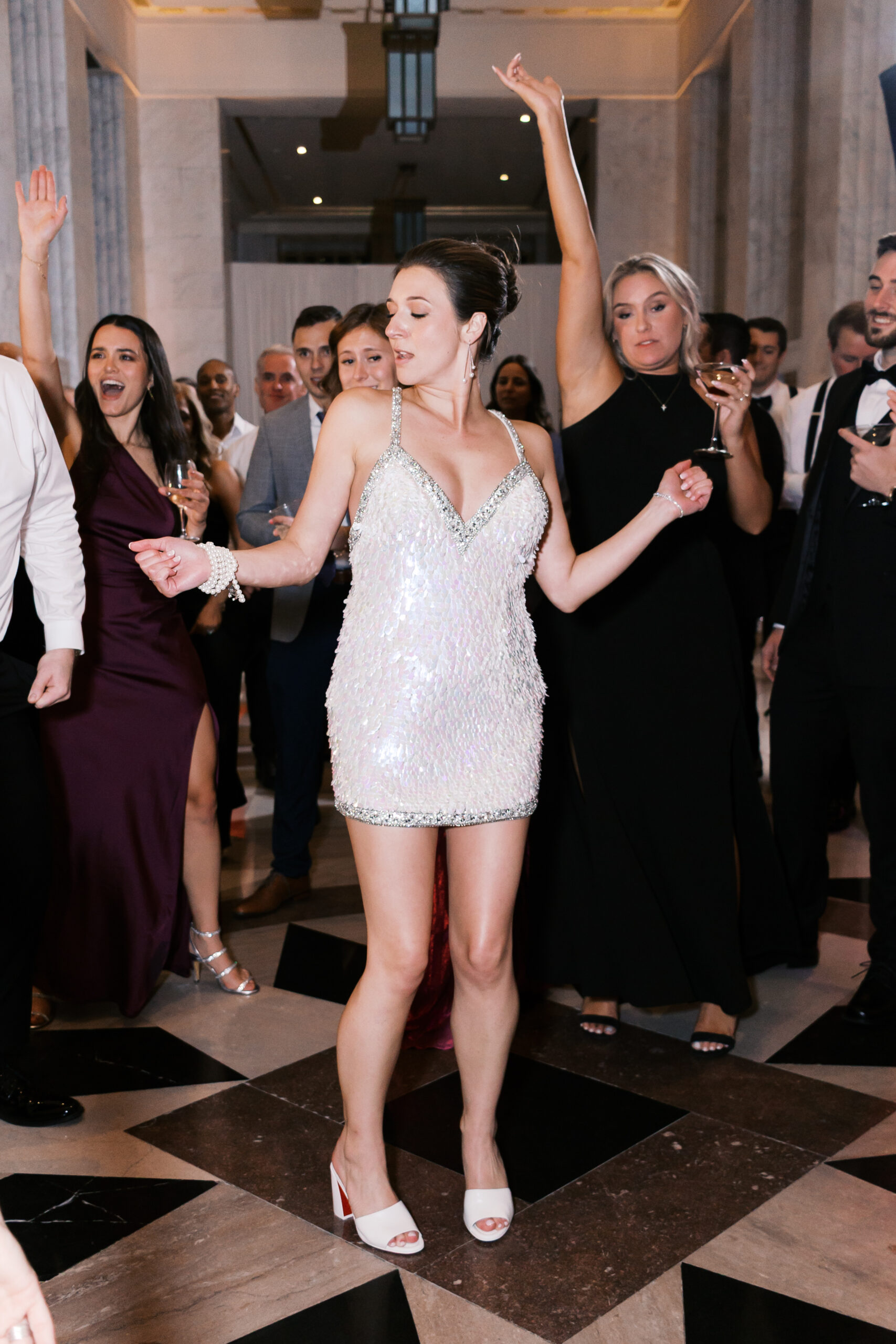 Bride dancing in a short sequined dress as friends and guests cheer in the background.