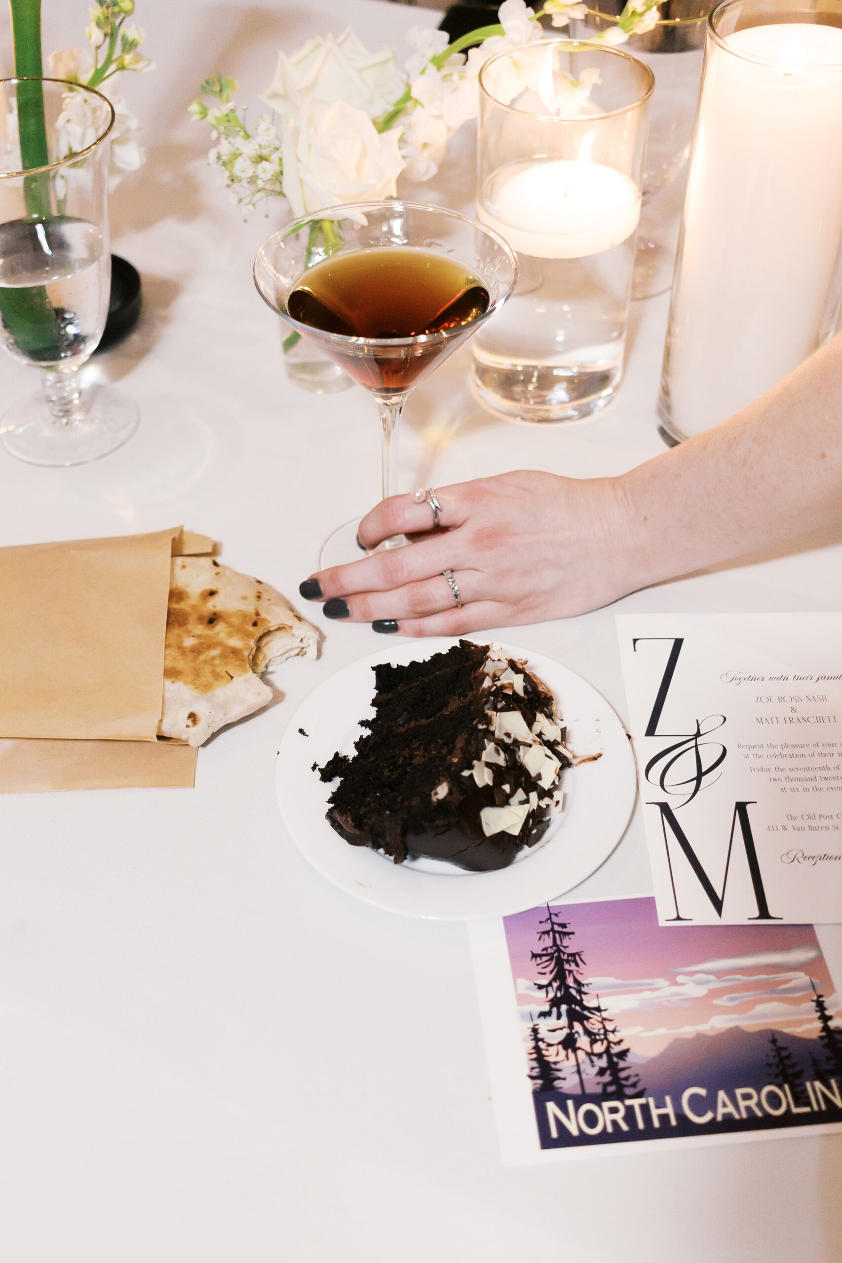 A hand holds an espresso martini in this reception flatlay of a taco bell chalupa, piece of wedding cake and the wedding invitation.