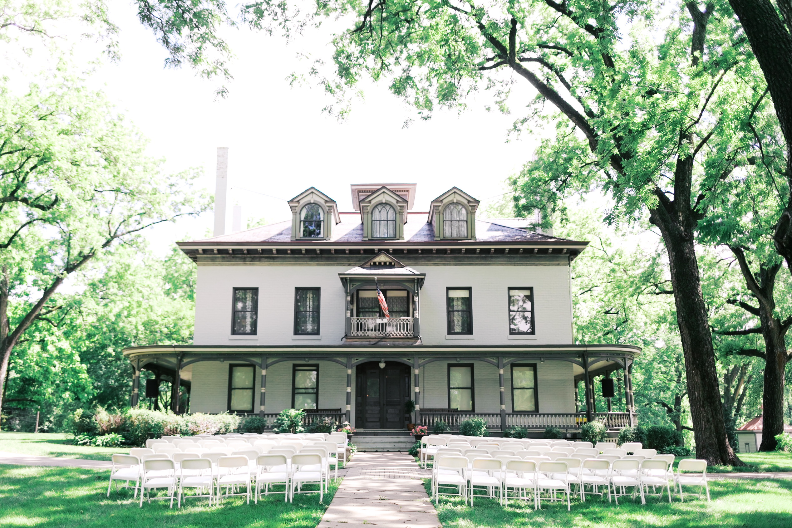 Exterior of a historic Victorian house with gray trim and wraparound porches, framed by leafy branches.