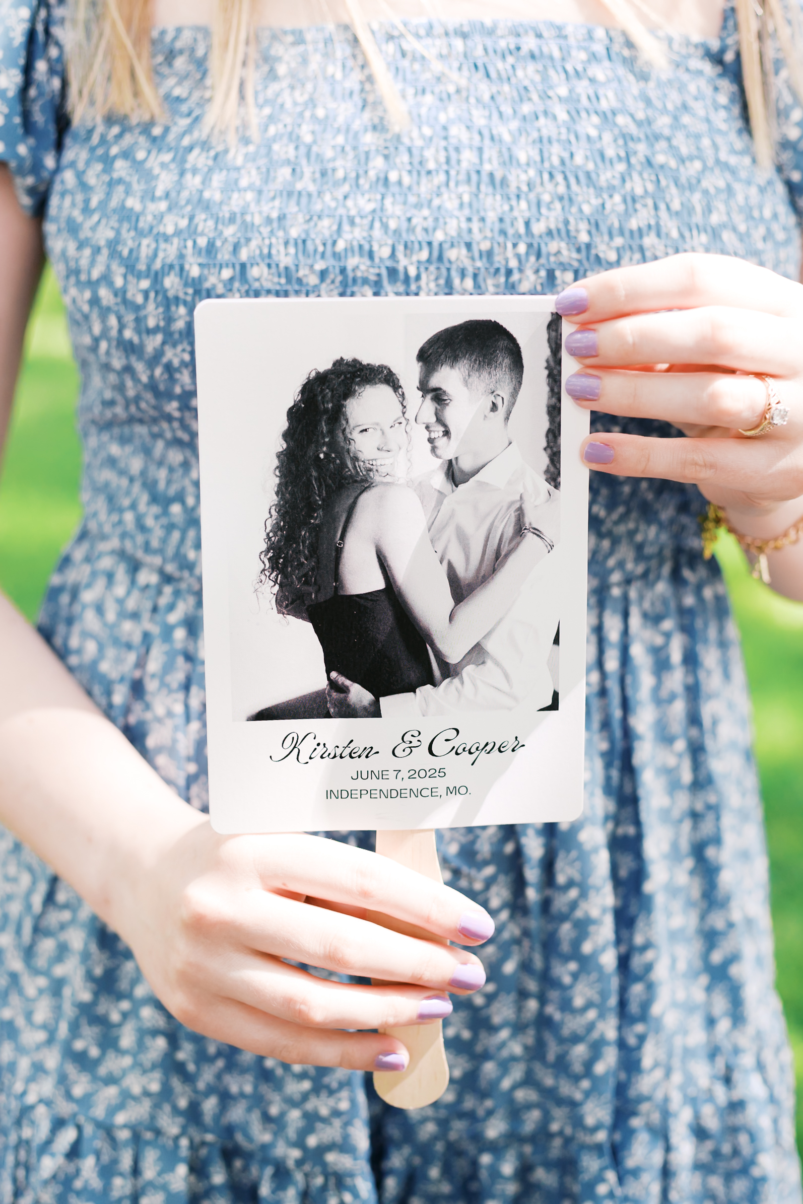 Woman in a blue dress holds a wedding fan printed with a black-and-white engagement photo and the couple’s names and date.