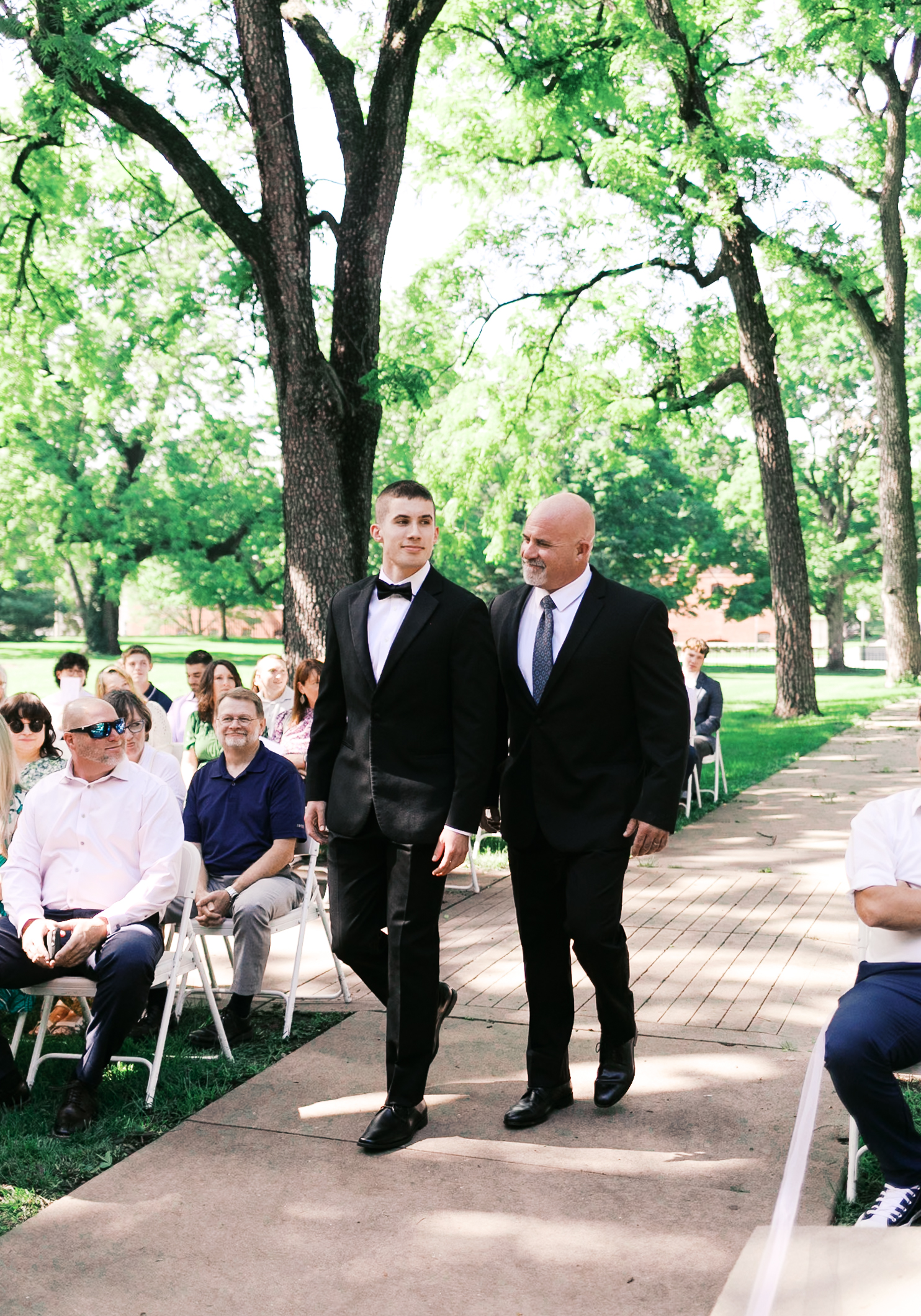Groom in a black tuxedo walks arm-in-arm with an older man during an outdoor ceremony, surrounded by seated guests.