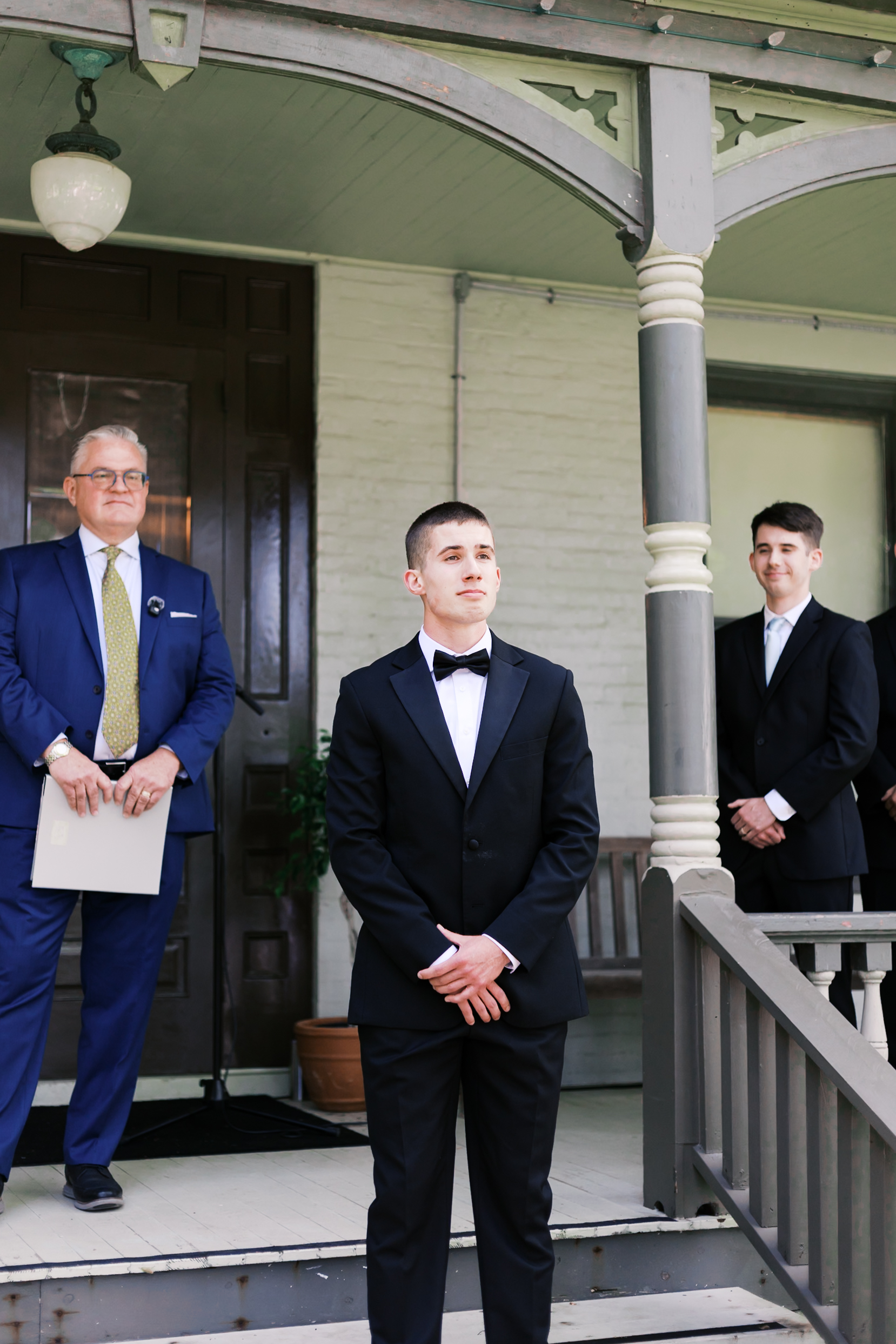 Groom in a black tuxedo stands with officiant and groomsmen on the porch steps, anticipating the ceremony.