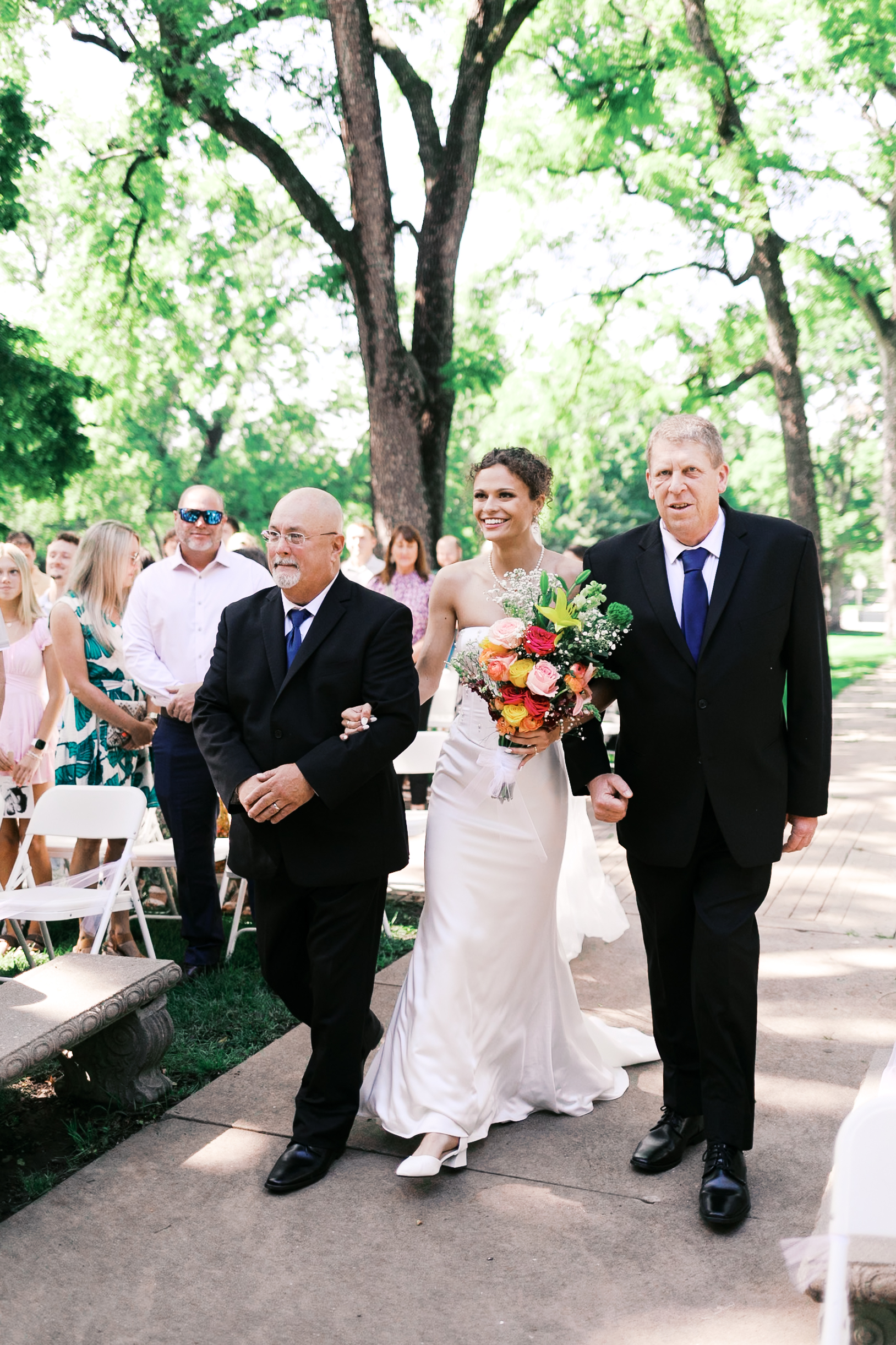 Bride in a white gown holding a colorful bouquet walks arm-in-arm with two men during an outdoor ceremony.