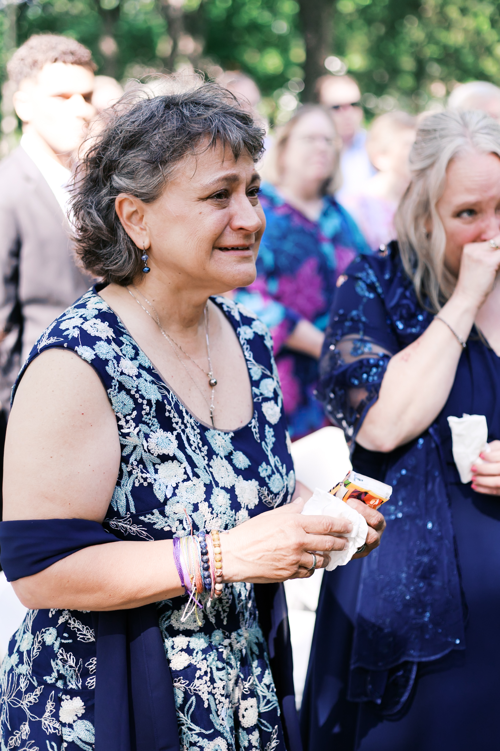 Close-up of woman in a navy floral dress holding tissues and a phone, visibly emotional during the ceremony.