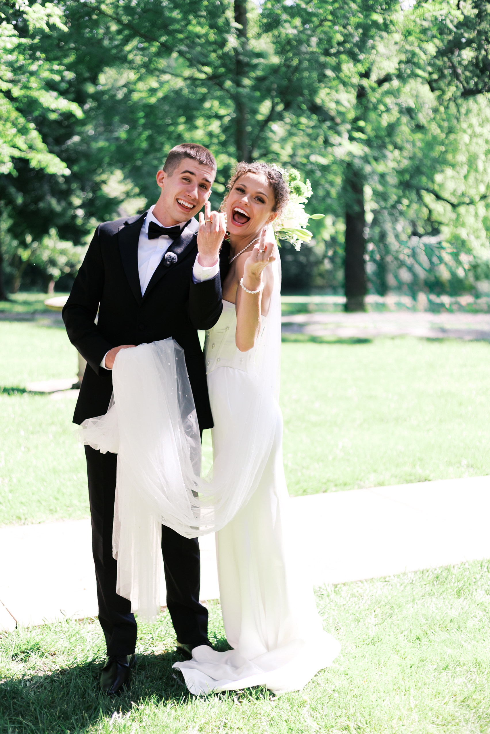 Bride and groom hold up their ring fingers and smile widely while showing off their wedding bands in a sunlit field.