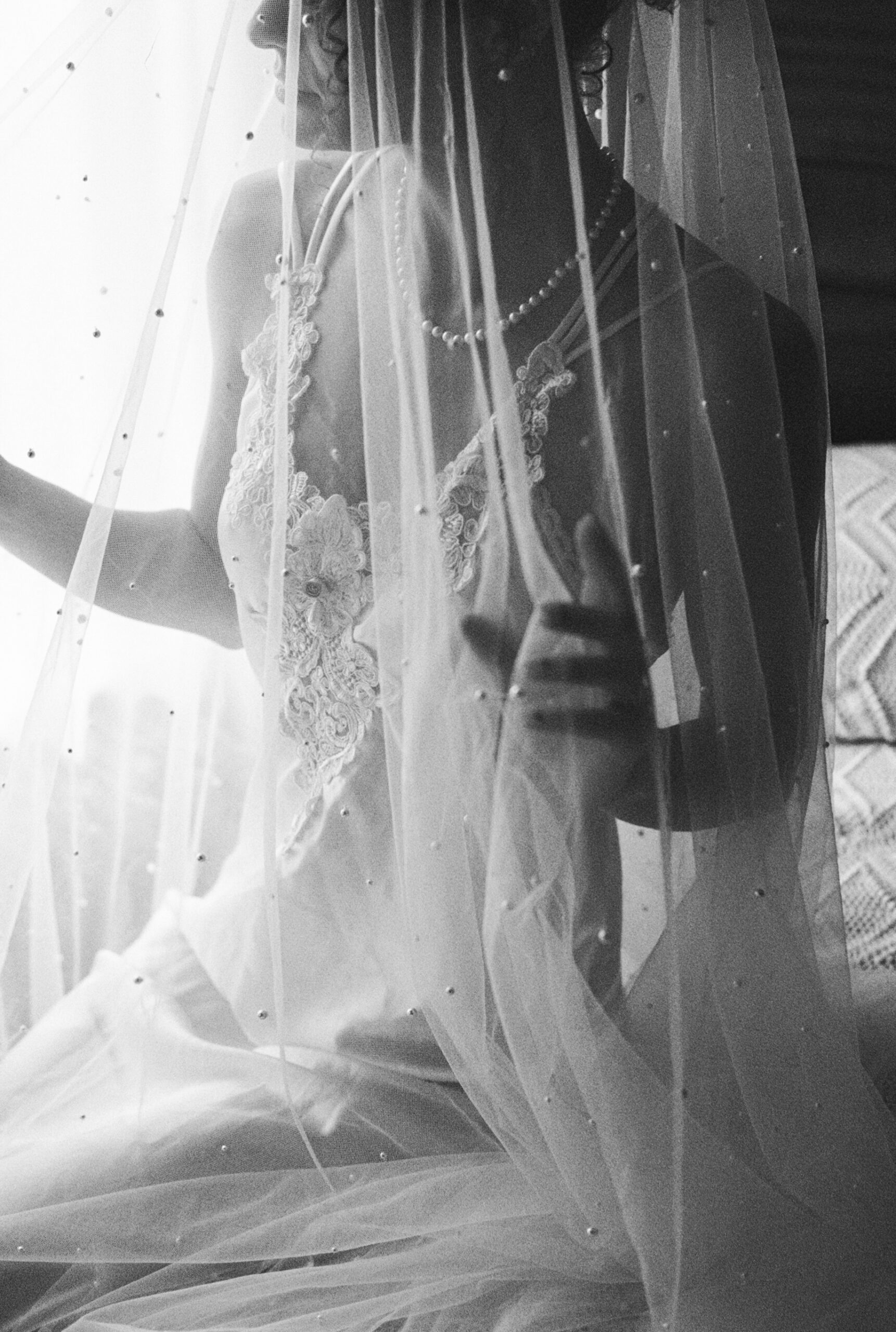 Grainy black and white close-up of a bride’s lace-trimmed slip and pearl necklace beneath a sheer veil with delicate dots.