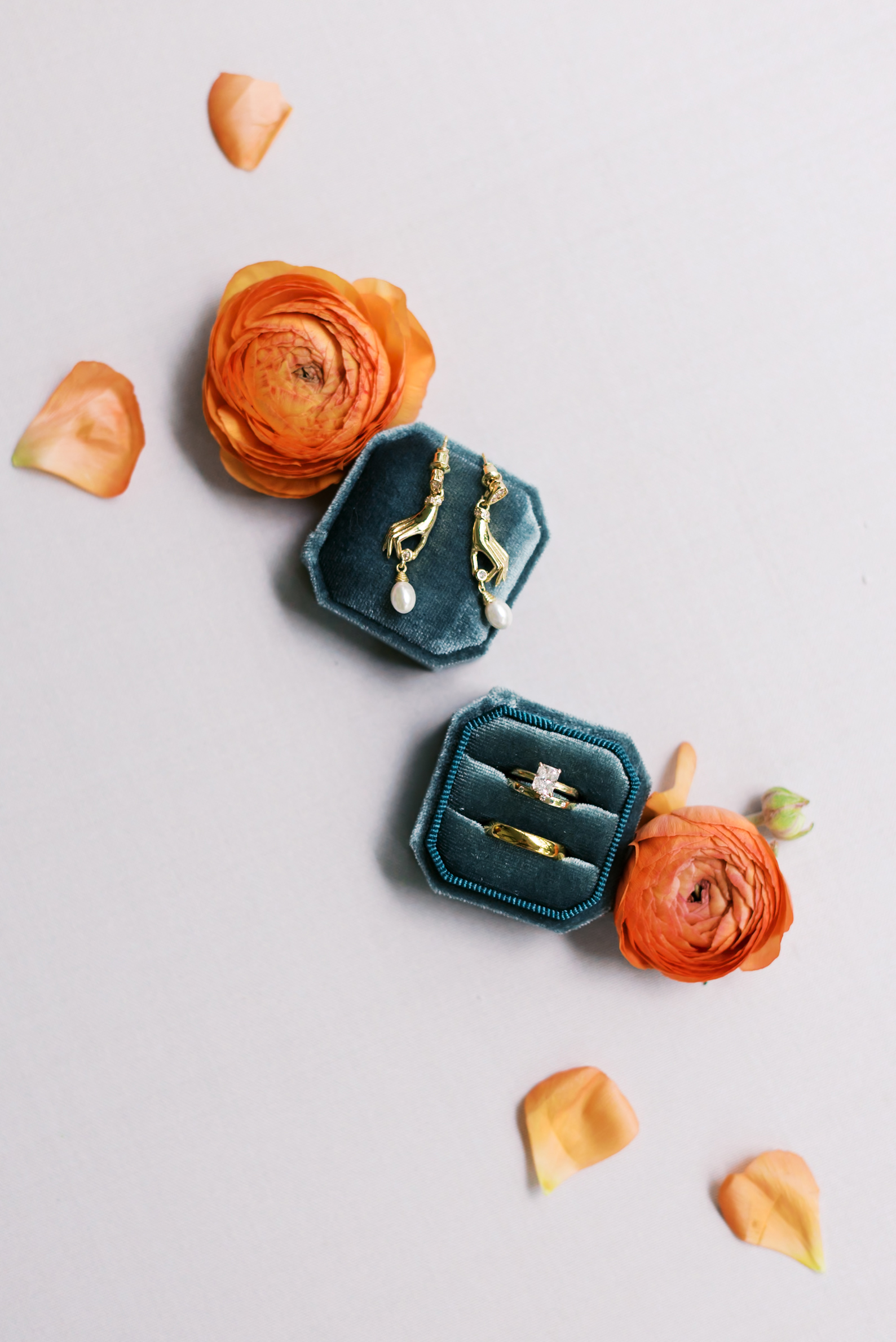 Flat lay of gold and pearl earrings beside velvet ring boxes and bright orange ranunculus flowers on a soft linen backdrop.