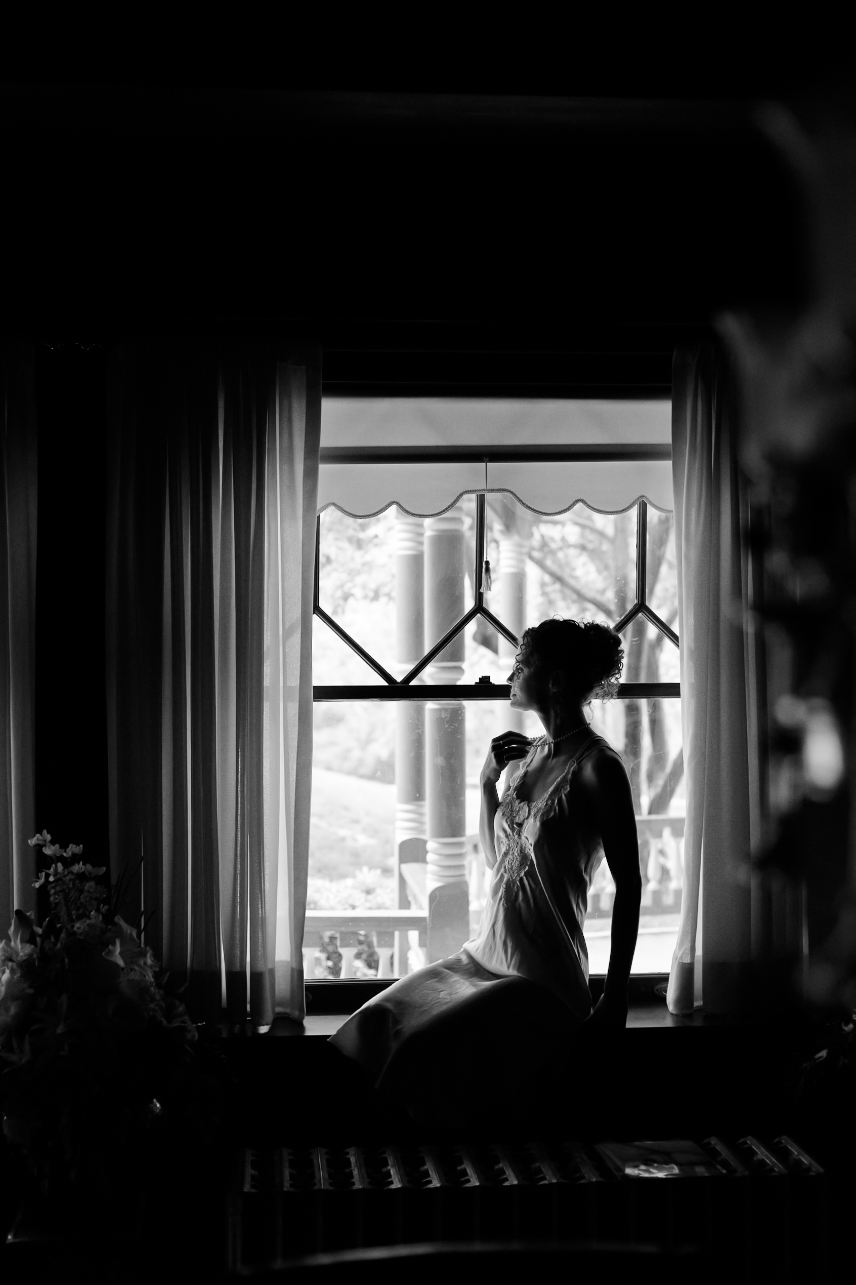 Black and white portrait of a bride sitting in a windowsill, softly lit by natural light through sheer curtains.
