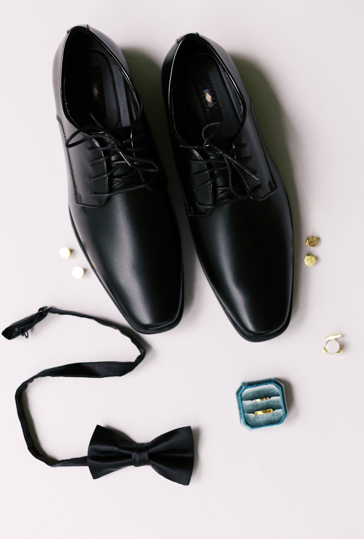 Groom’s black dress shoes, bow tie, cufflinks, and wedding bands arranged neatly with velvet ring box on a light background.