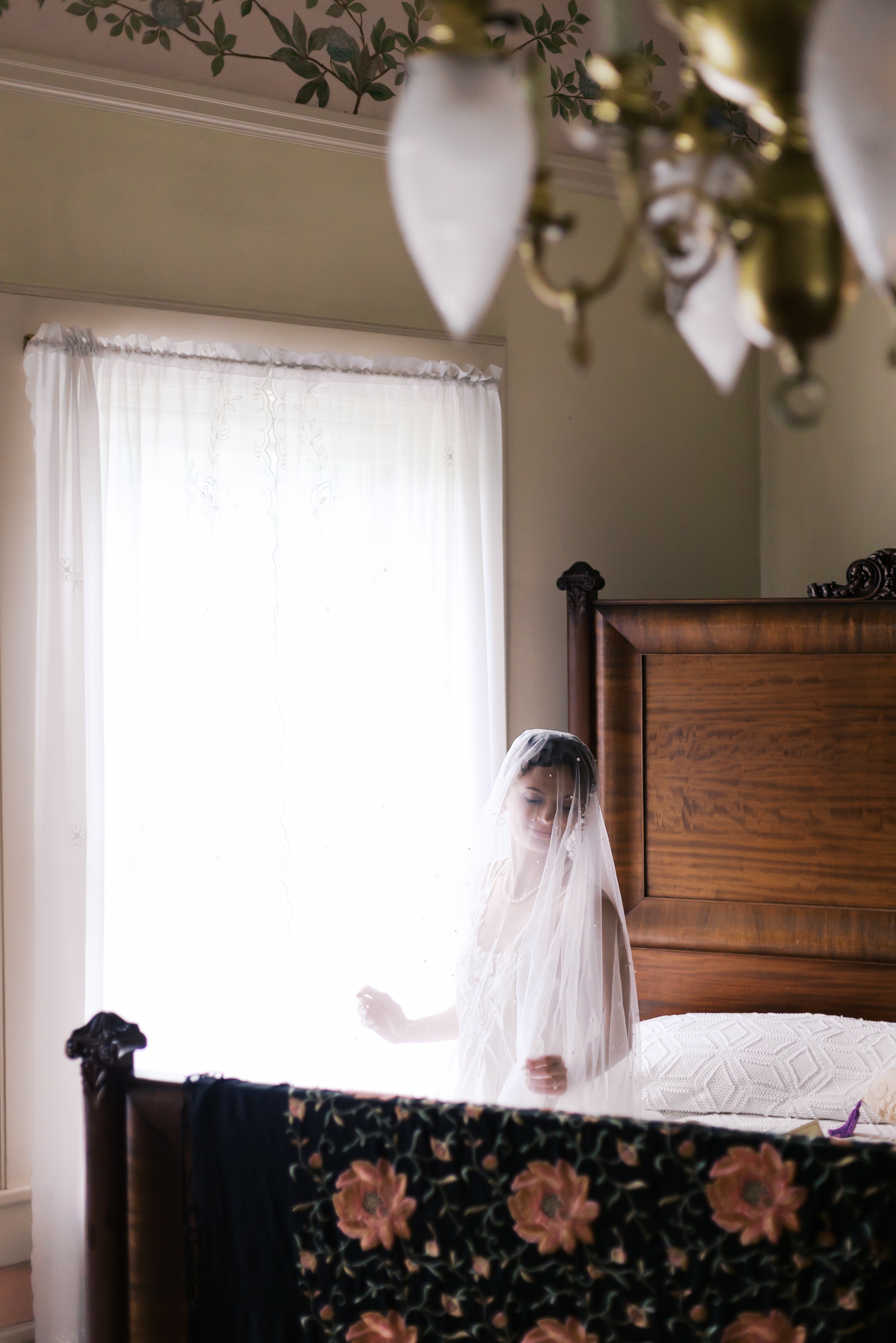 Bride in a vintage bedroom, softly backlit by window light, wearing a long sheer veil and a lace-trimmed slip dress.