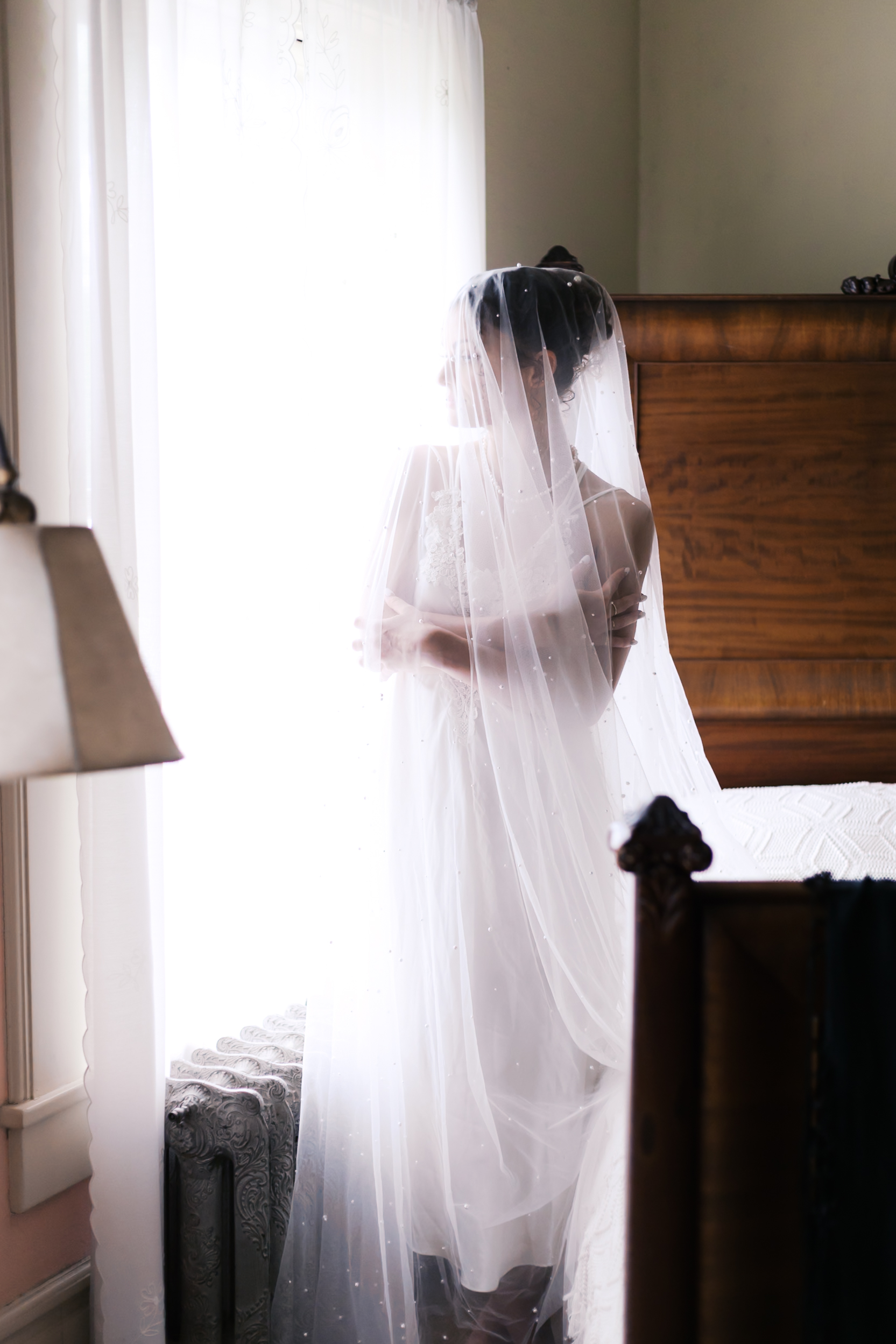 Bride wrapped in a sheer veil, standing beside a bright window, arms gently folded across her chest.